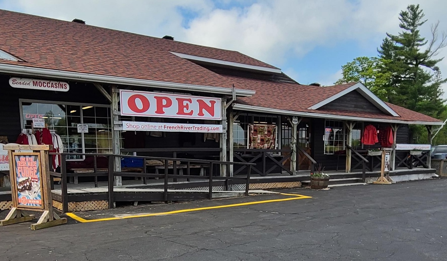 The exterior of the French River Trading Post; a long wooden building with a brown roof and signs advertising fresh fudge, moccassins, and other gifts. 