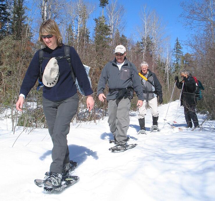 4 smiling snowshoers move down a snowy forest trail on a sunny winter day.