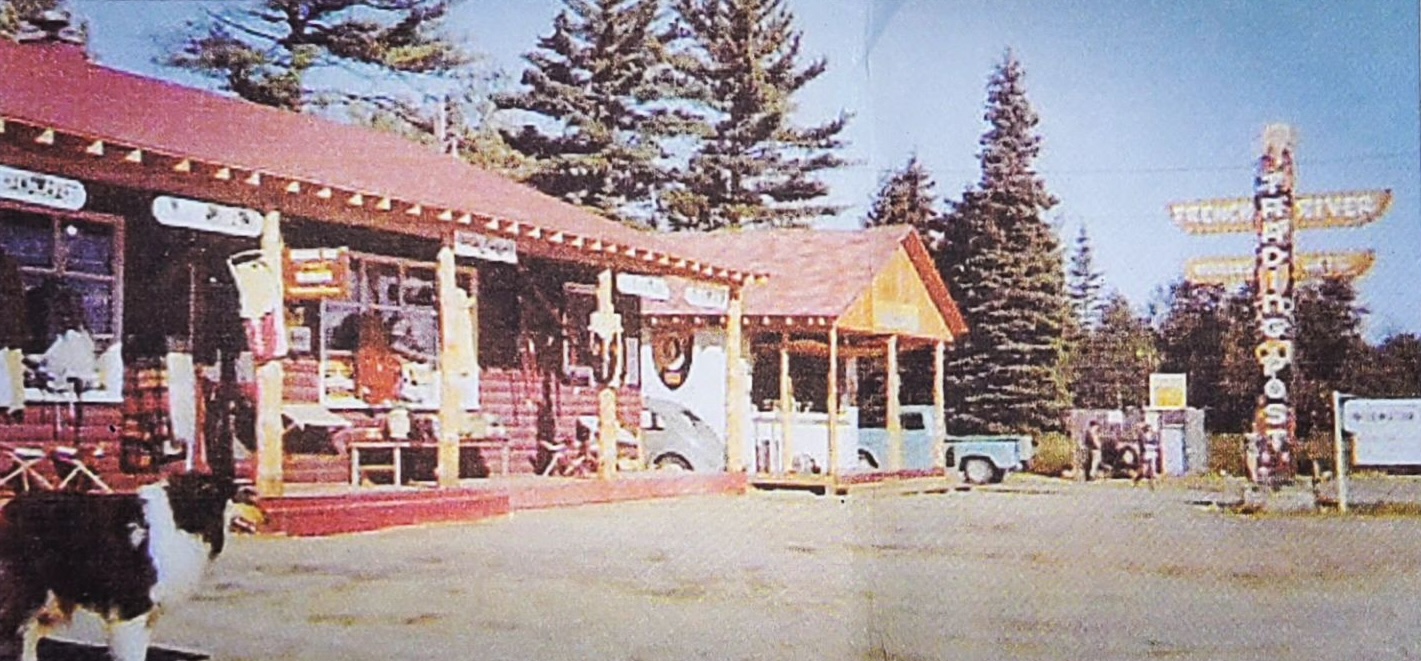 An old coloured photograph of the French RIver Trading Post in its early days, with lots of gifts and carved wooden statues out front on a sunny summer day. 