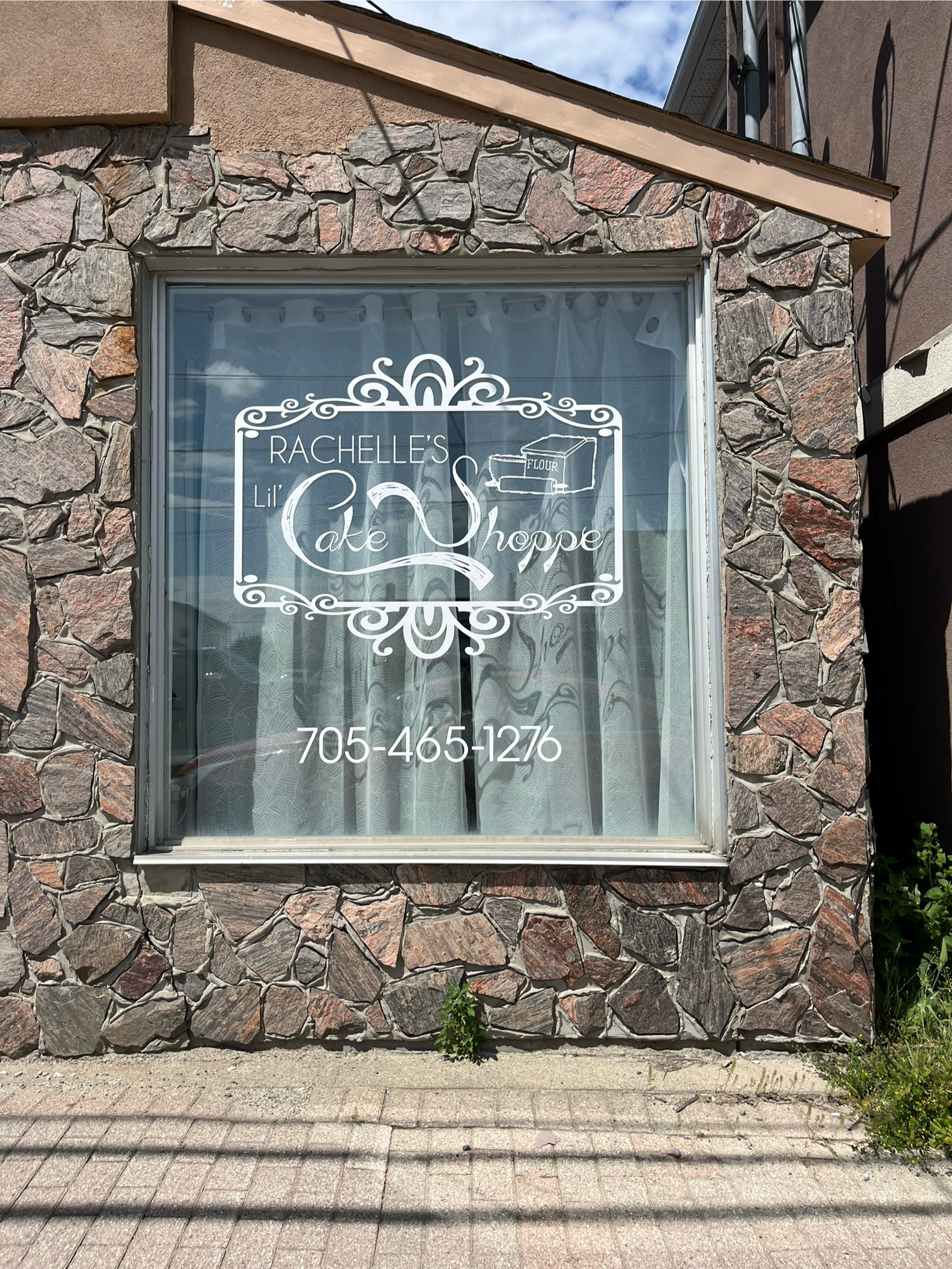 a close-up of a brick building with a window in the center with the Rachelle's Lil' Cake shop logo and phone number displayed on the window and curtains behind it