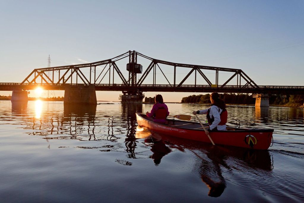 two paddlers in a canoe approach the LIttle Current Swing Bridge on sparkling, glassy water at sunset. 