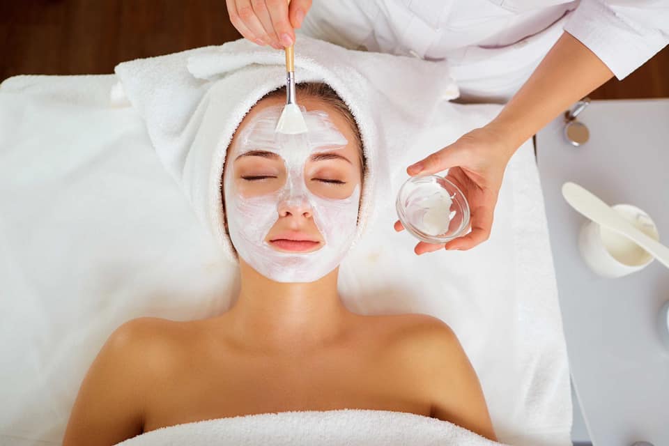 a woman wrapped in white towels laying on a spa table relaxes with closed eyes as an esthetician applies a facial mask with a brush.