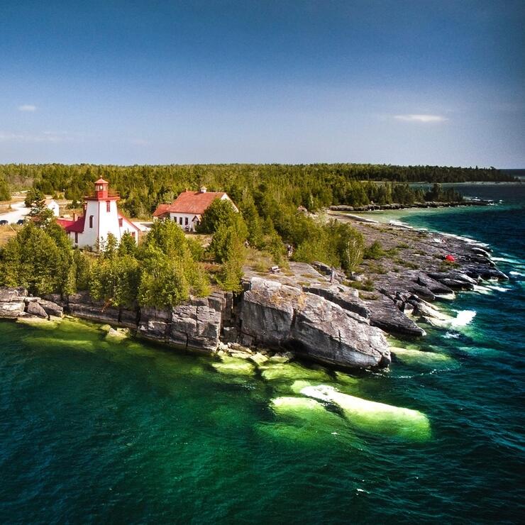 The Mississagi Lighthouse, standing on the rocky and forested shore of Meldrum Bay, surrounded in clear turquoise water under a bright blue sky.