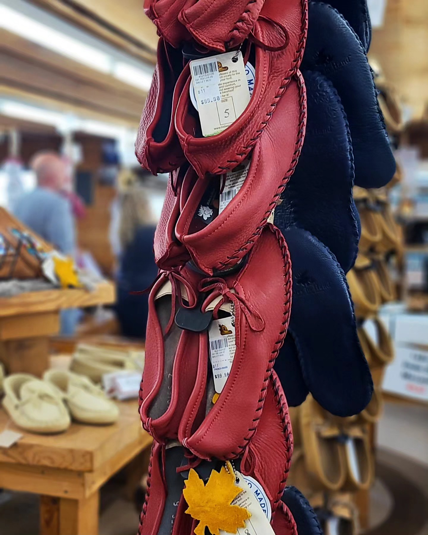 Leather moccassins in a variety of colours on display in the French River Trading Post. Shoppers are perusing the goods on the wooden display tables in the background. 