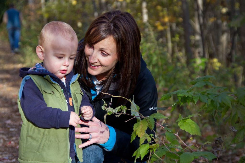 A mother smiles as she speaks to her young child, who is looking closely at a leaf in a forest.