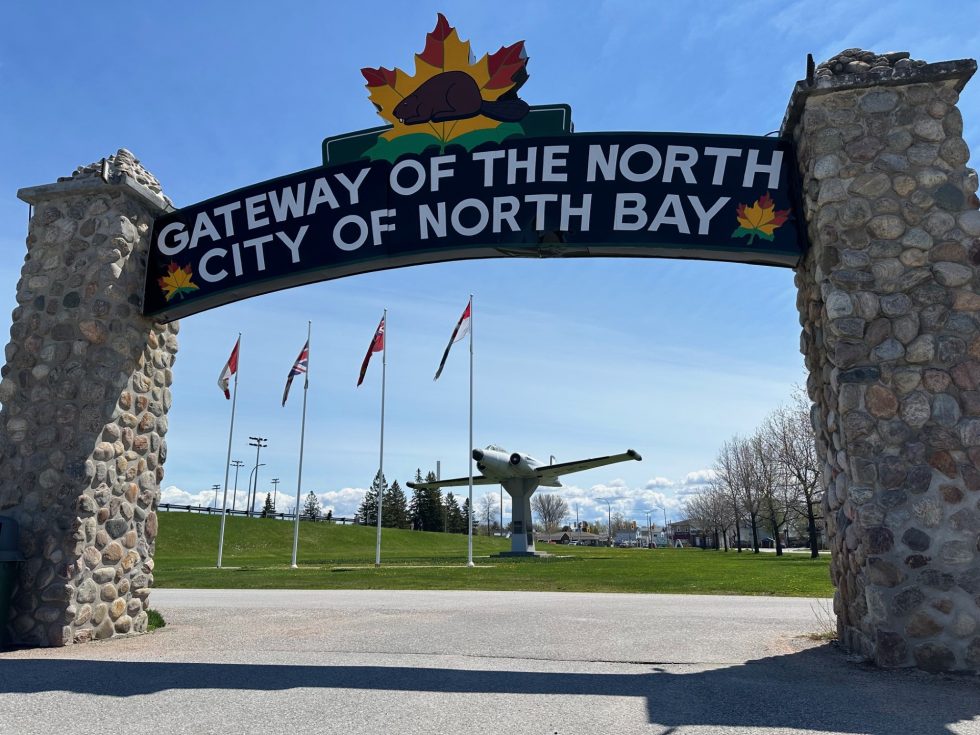 A stone and wooden archway that says Gateway of the North City of North Bay. Through the arch, a fighter aircraft on a pedestal is visible. 