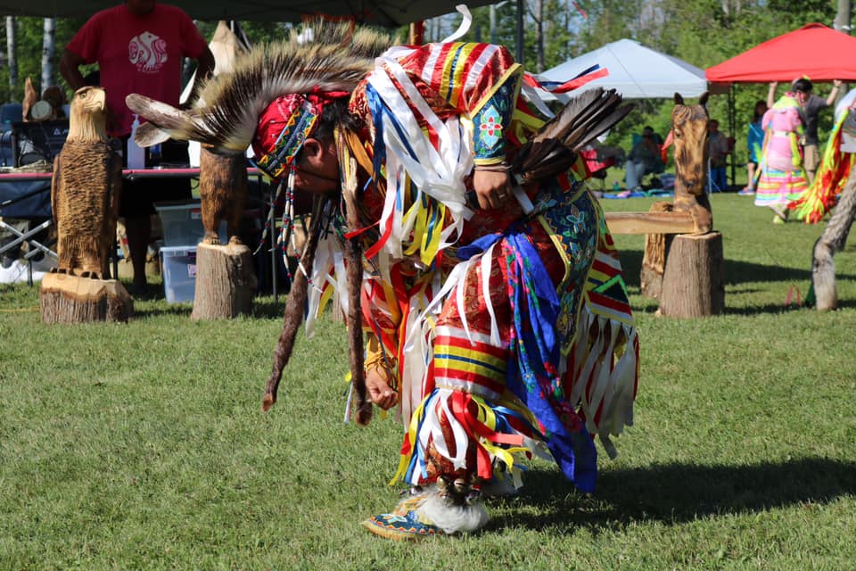 A man in beautiful brightly coloured and feathered regalia dances in the grass at the Sheguiandah Annual Jiingtamok.