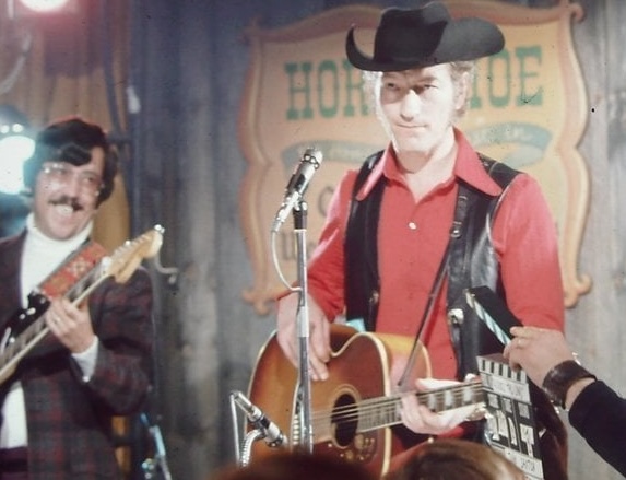 A young Stompin' Tom Connors in a red shirt, black cowboy hat and black leather vest at a small concert in a wooden-walled saloon. One of his bandmates is looking at him and smiling as though he's just told a joke. 