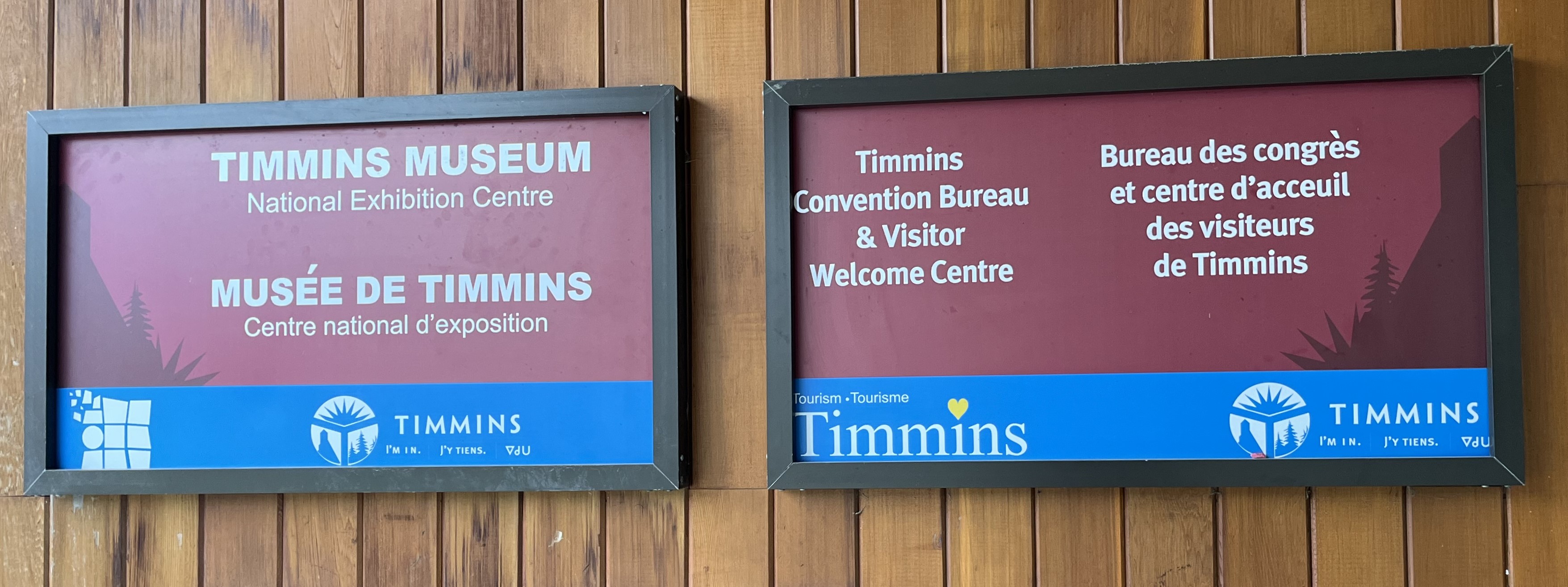 a close-up on the Timmins Museum sign on the left and the Tourism Timmins sign on the right which hangs from the outside of the Timmins Museum building