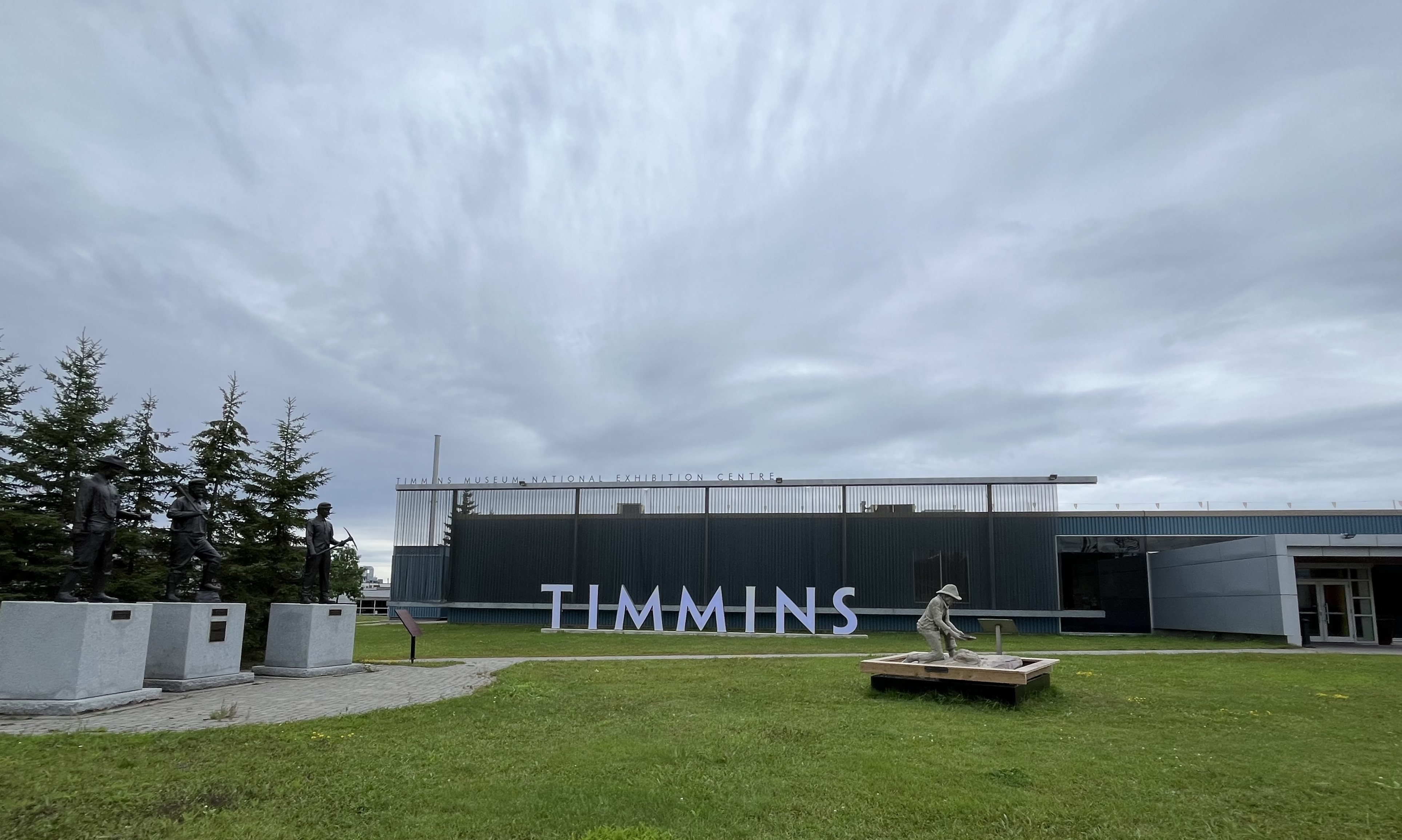 A landscape view of the right side of the exterior of the Timmins Museum. On the left hand side are the statues and the is a statue on the right hand side as well with the blue glowing Timmins sign in the distance in the center of the image.