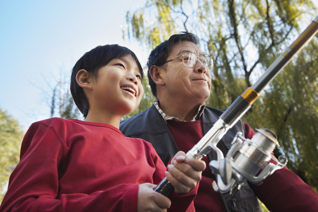 an older man with glasses and a young boy with a fishing rod, both smiling as they fish together on a summer day. 