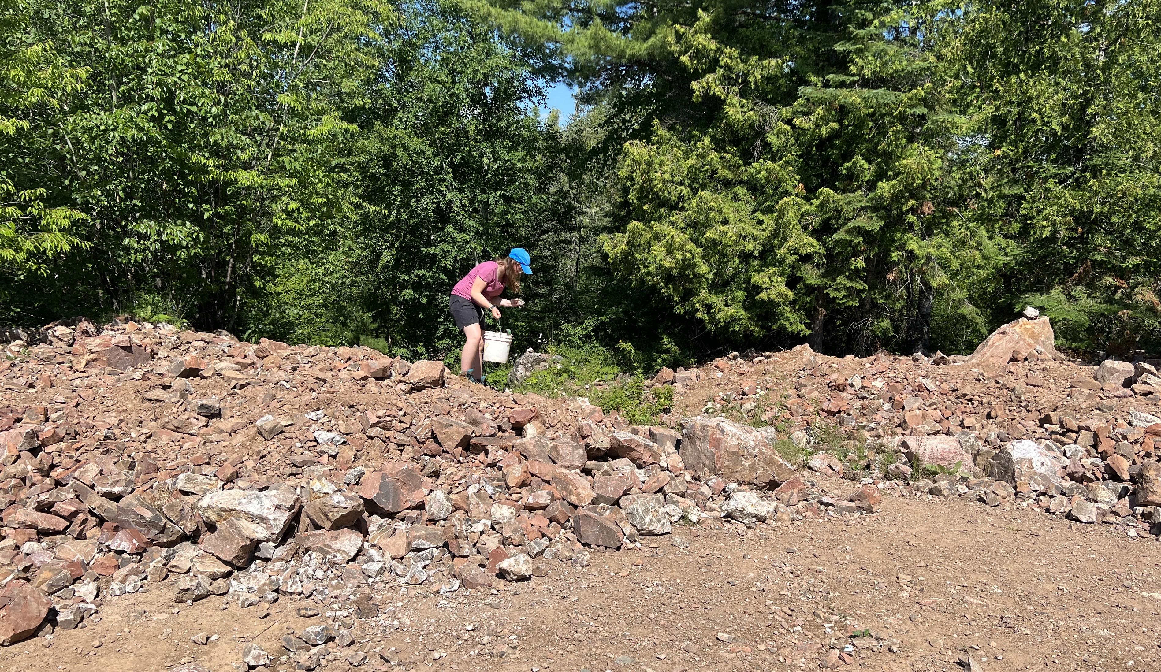 a child holding a bucket looking through a broad rocky mine site for amethyst.