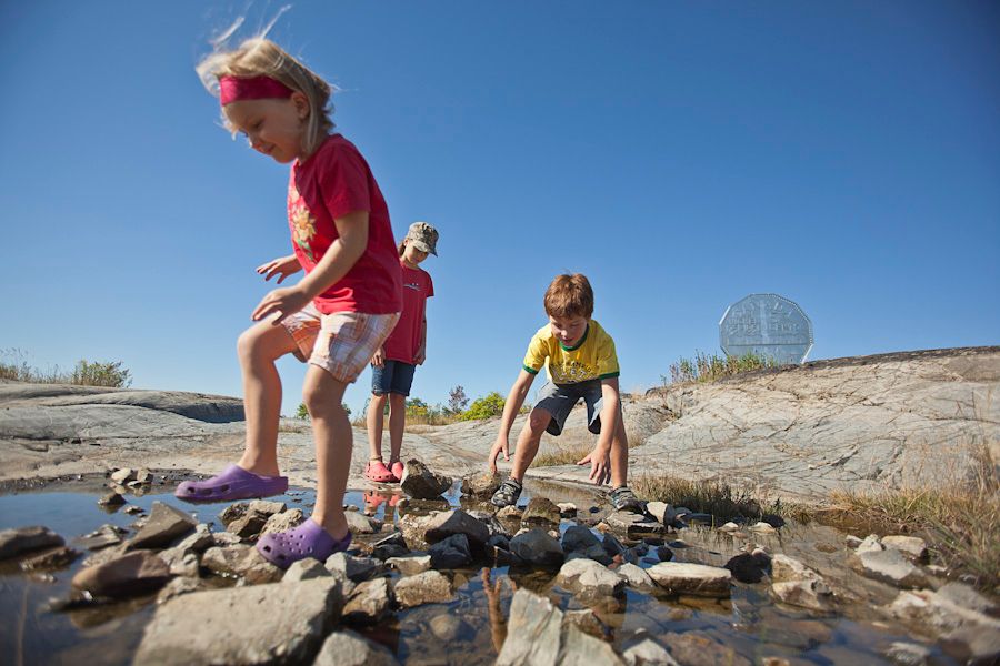 Children playing amidst some rocks and water on a summer day with the Big Nickel in the background.