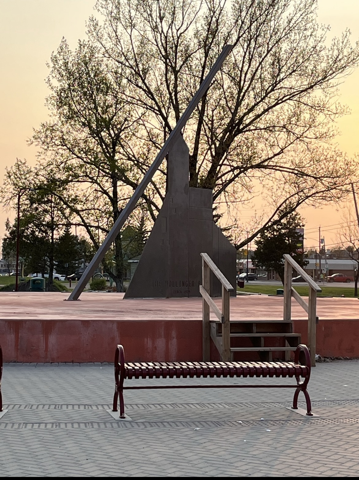 A Close-up of the Hollinger Park Sundial which stands on a red platform. There is a red bench in front of it and a large tree without its leaves behind. There is also a sunset as the background of the image and various other trees in the distance. 