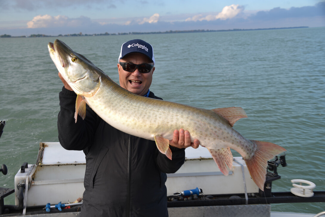 A smiling man holding up a muskie that's nearly as long as he is in front of a lake.