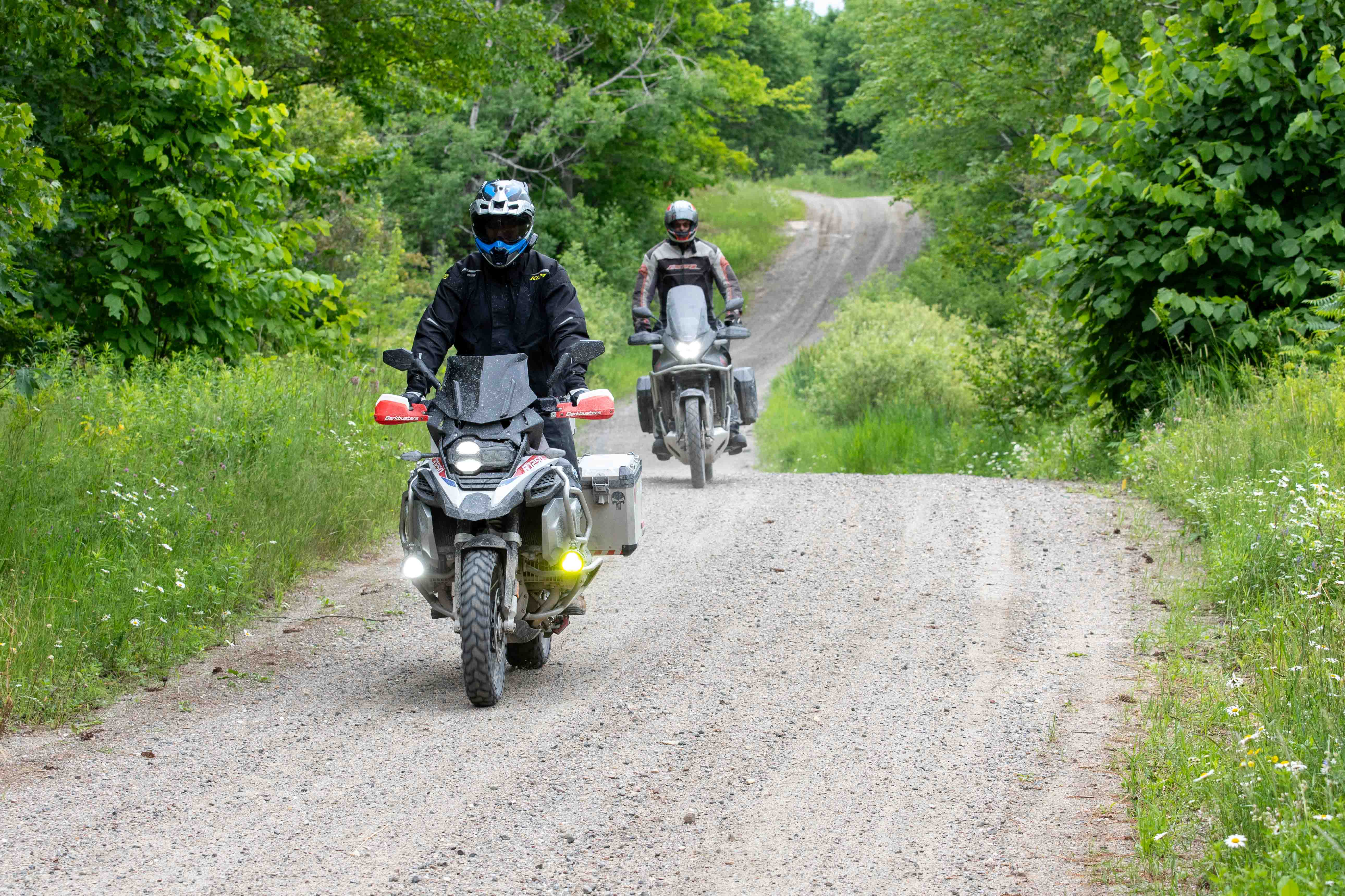 2 motorcyclists ride down a narrow, winding gravel road through lush green forest.