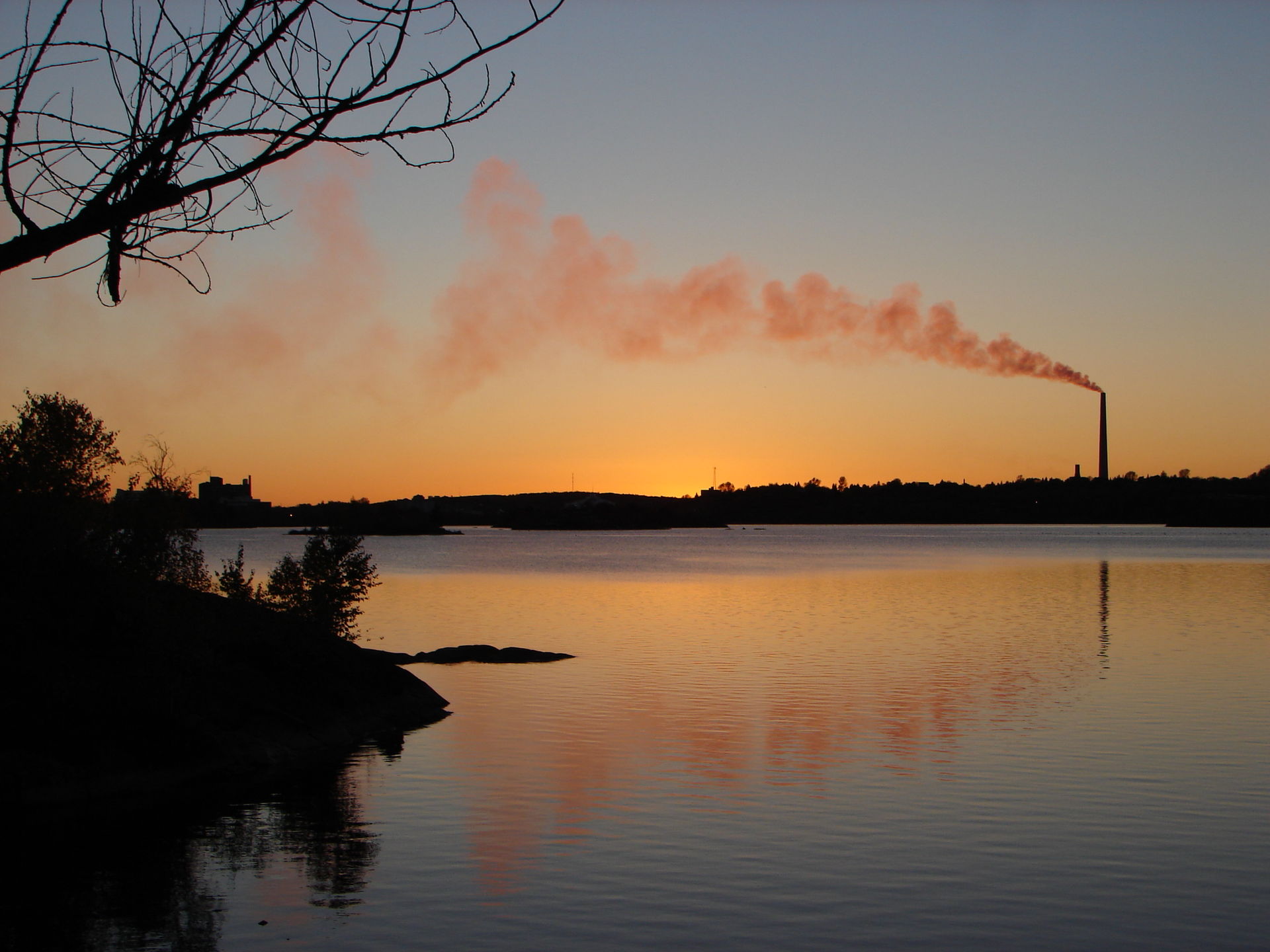 A calm, glassy lake at sunset, with the Sudbury Superstack silhouetted on the far bank, a long trail of smoke blowing sideways from its top in the wind.