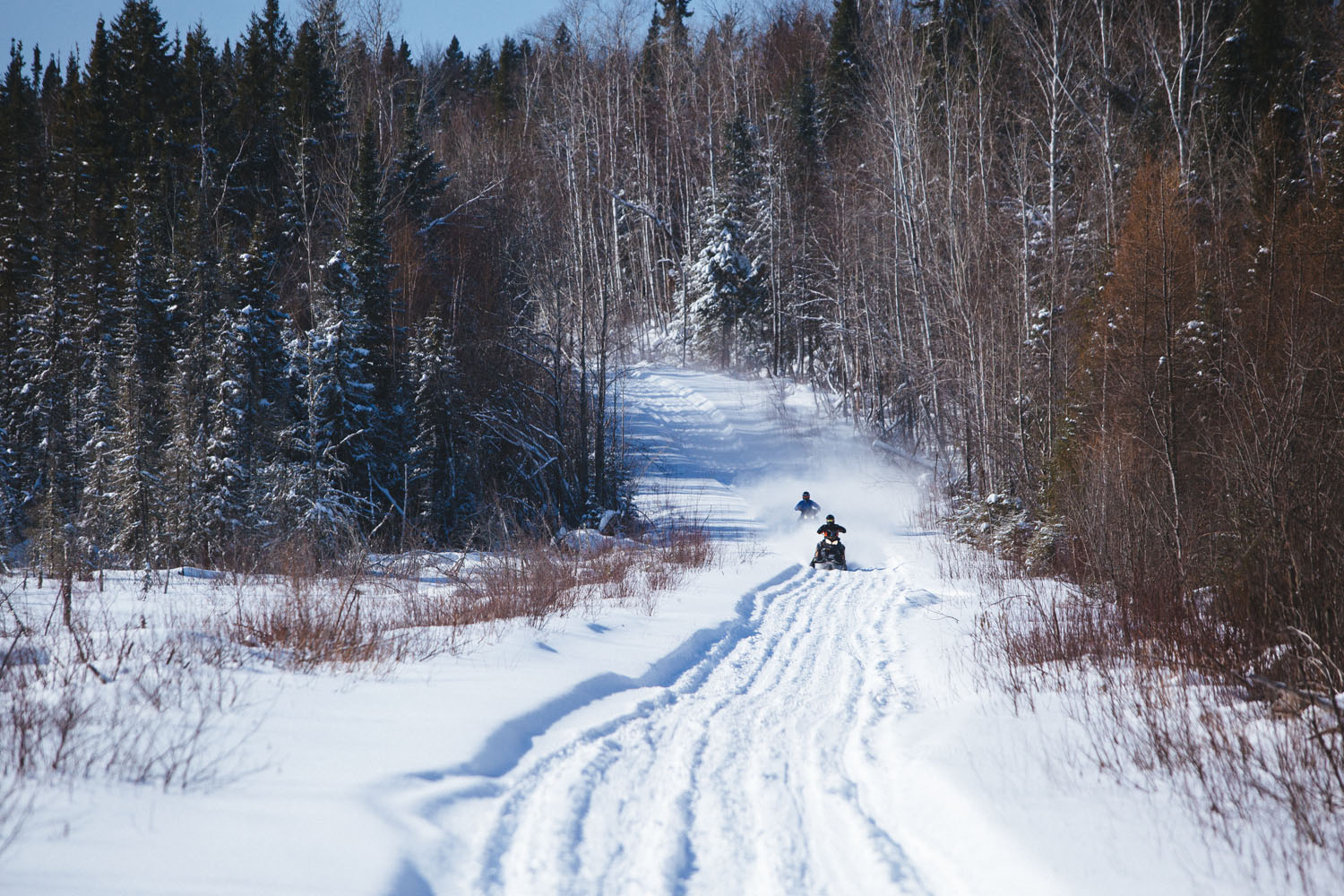 A landscape view of a snowmobile trail with 2 people in the distance who are snowmobiling along it