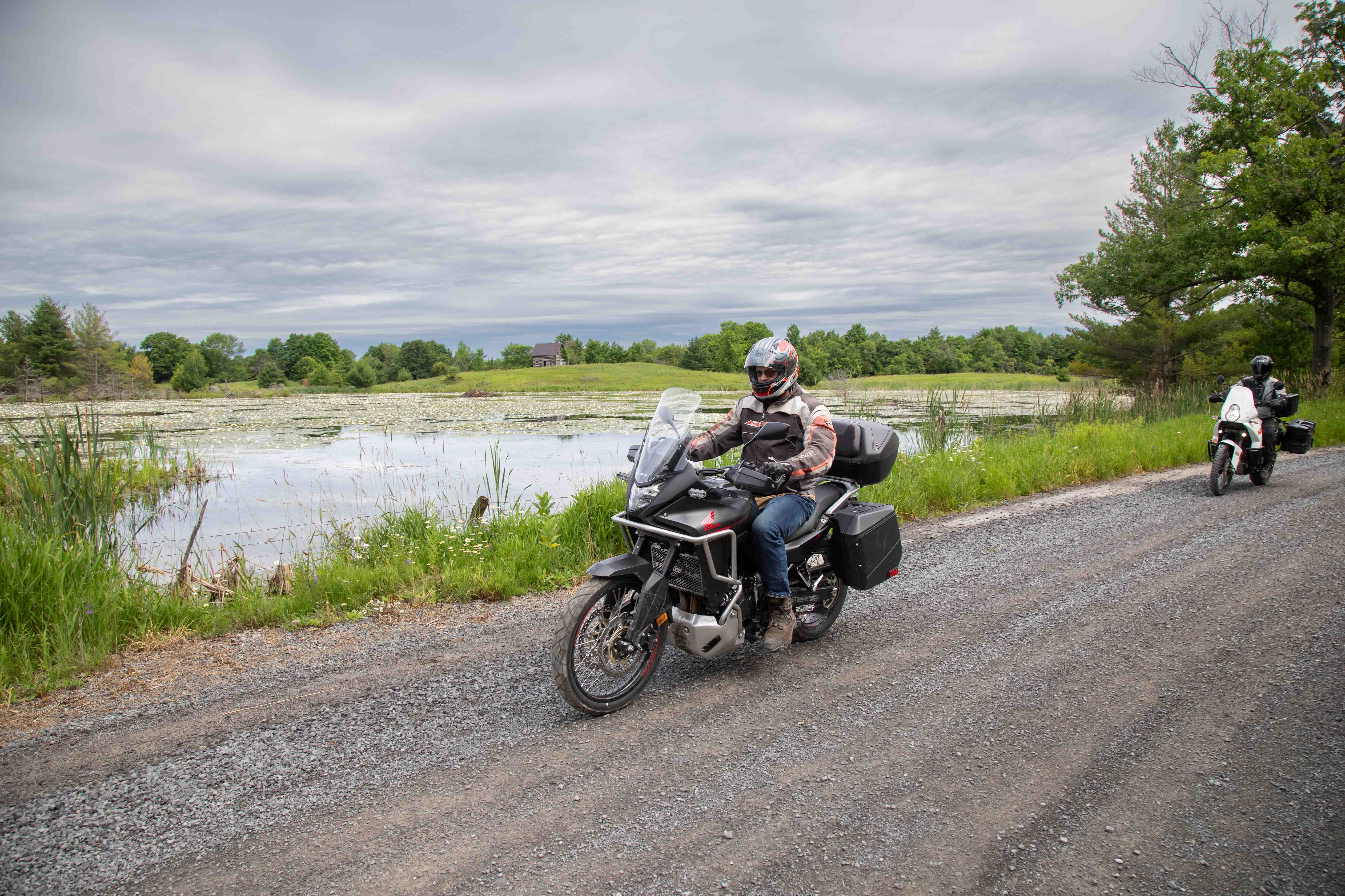 two motorcyclists ride down a gravel road past a shining pond in a forested area. 