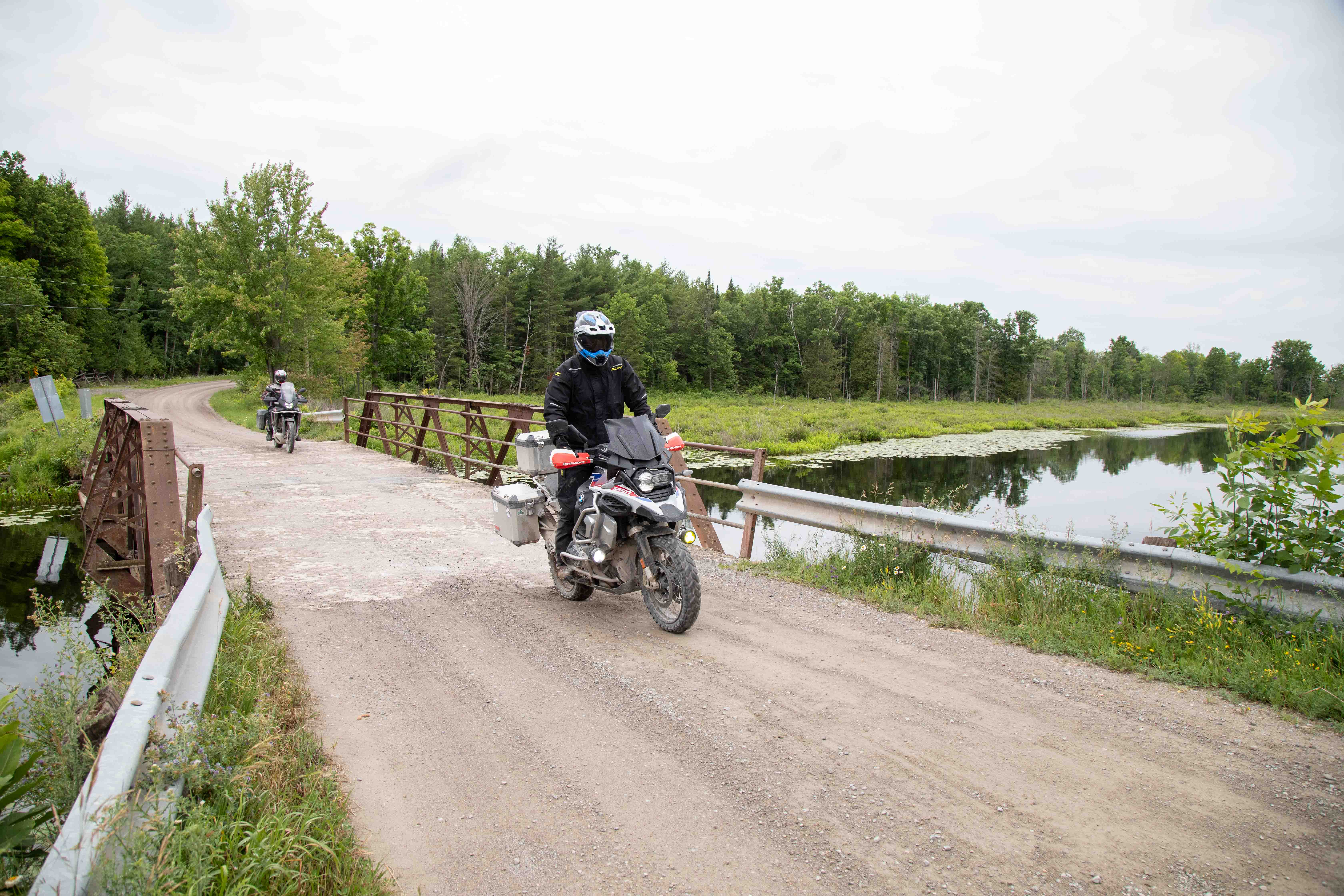 two motorcyclists ride across a small, pretty wooden bridge over a marsh surrounded by lush green forest and farm land. 