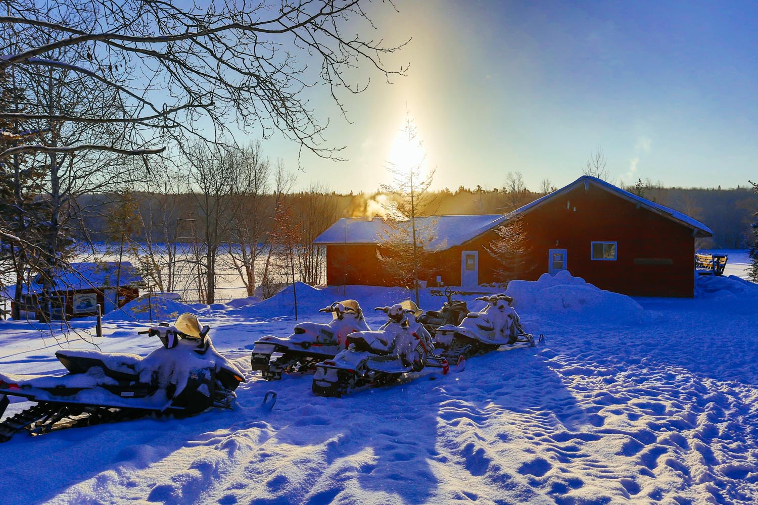 5 snowmobiles are parked in front of a cabin and are covered with snow. The sun is rising in the background and there are trees on the left-hand side including a smaller cabin in the distance. There is a lake behind the cabin