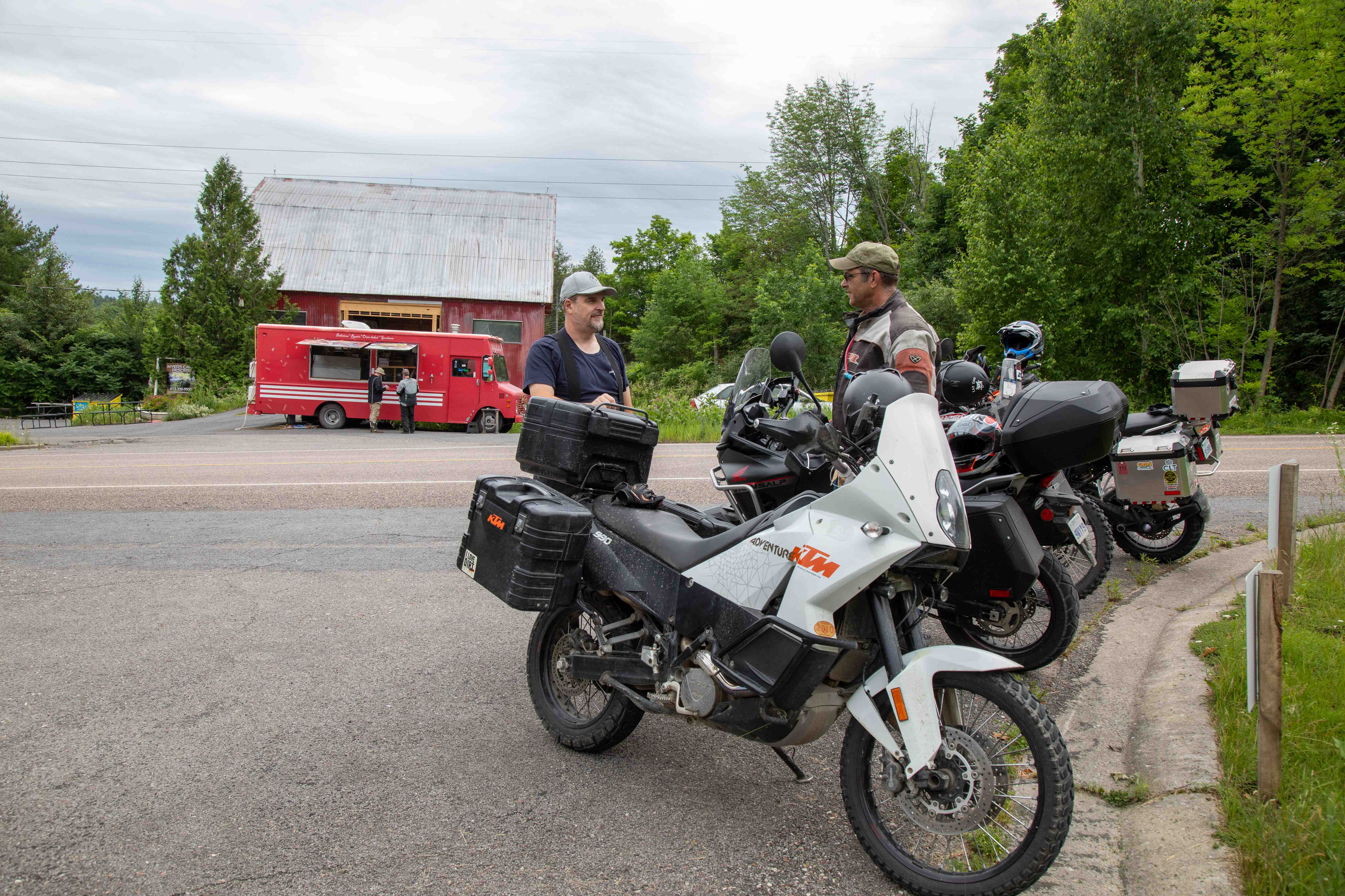 4 motorcyclists parked in a lot surrounded by forest, a red food truck and large red barn in the background. 