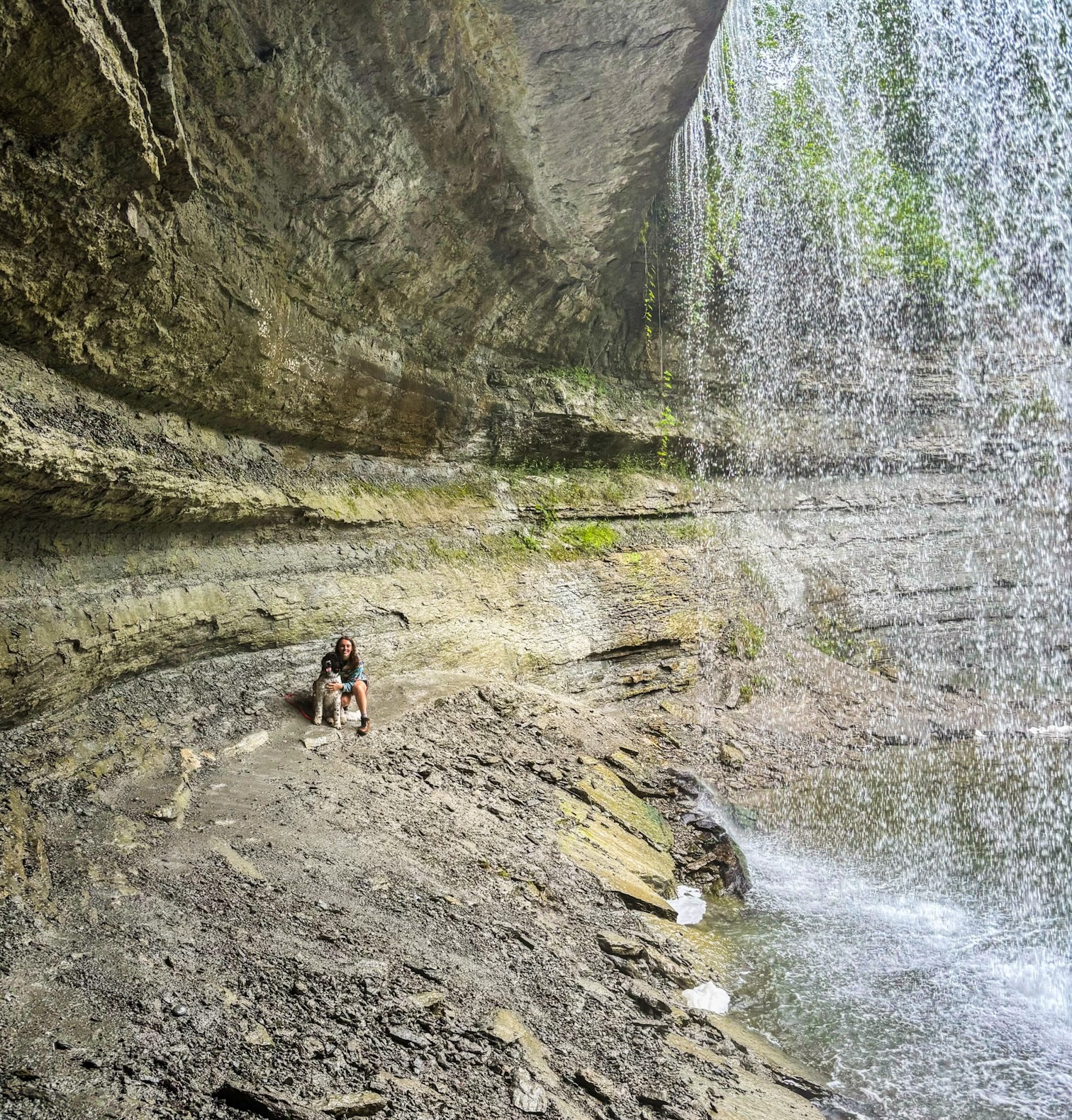 A smiling woman and her dog sitting on the large, mossy stone area behind the curtain of water of Bridal Veil Falls.