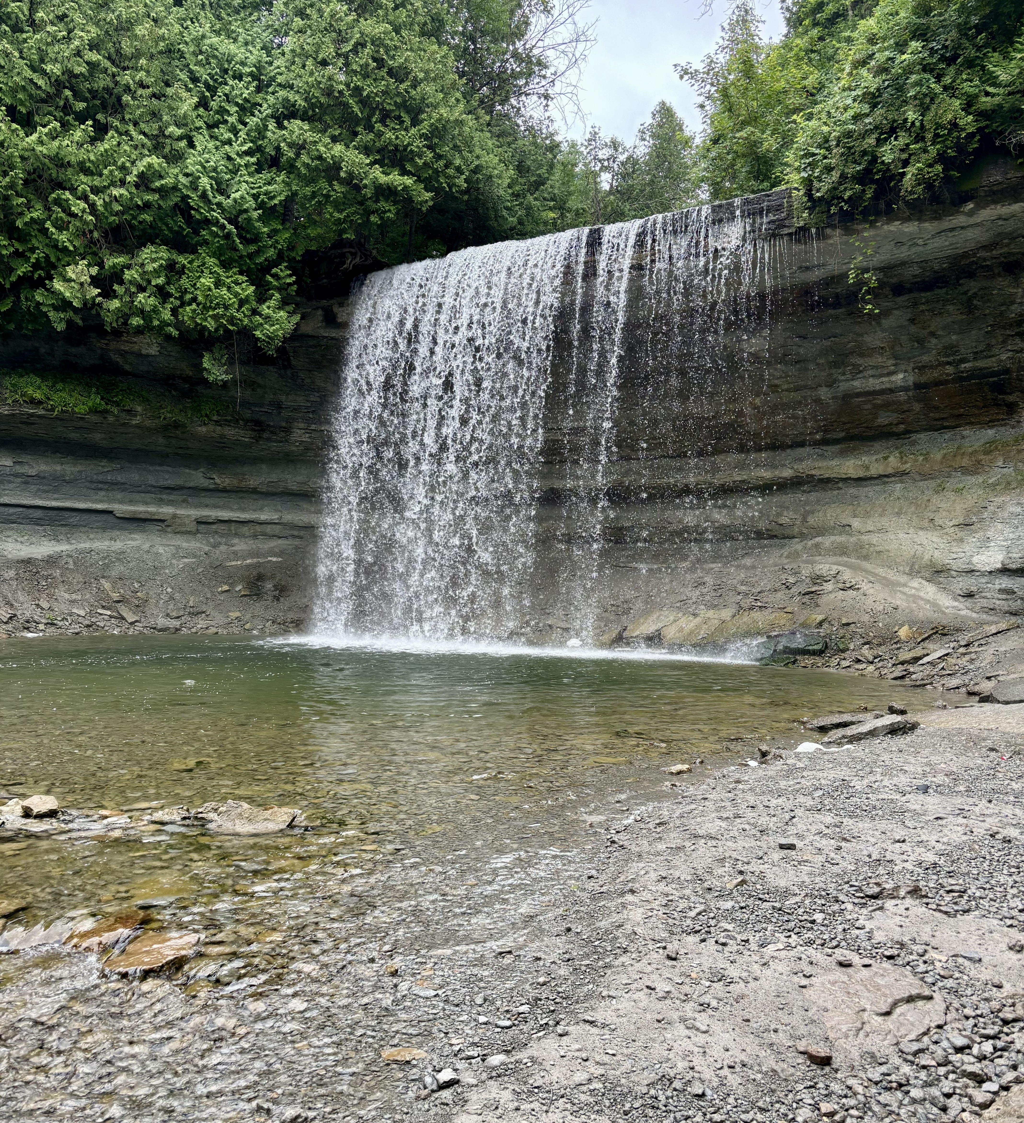 Bridal Veil Falls from the front; a wide gentle falls that plunges off a forested stone cliff into a calm rocky pool below. 