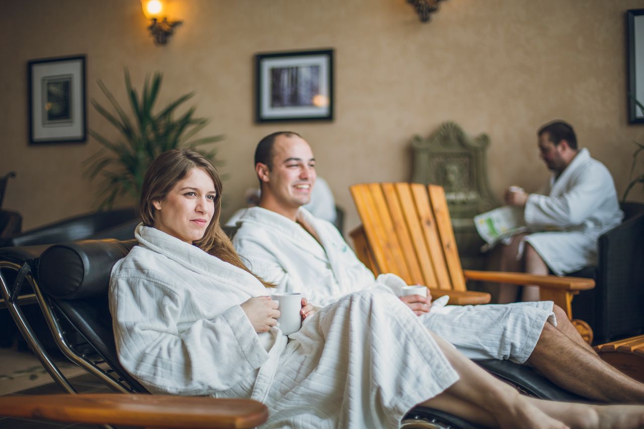 Two smiling people lounge in spa chairs while wearing white robes at Cedar Meadows Spa.