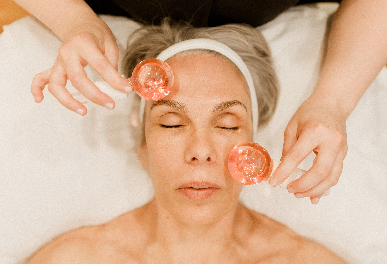 A close-up of a woman's face. Her eyes are closed and hands are shown holding two round pink objects onto either side of her face