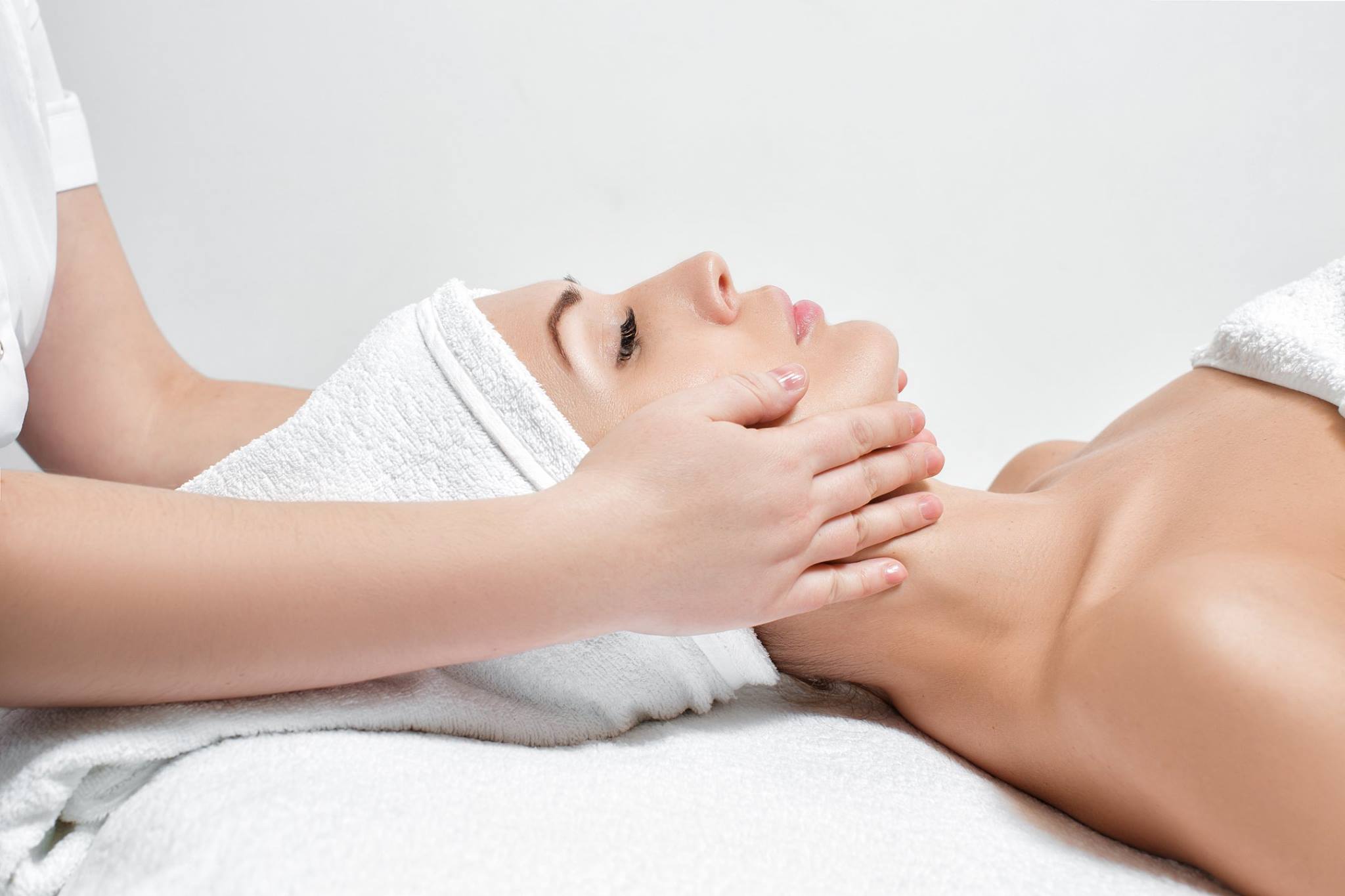 A side profile view of a persons head laying down with their eyes closed and a towel on their head as well as the esthetician's hands which are message their face