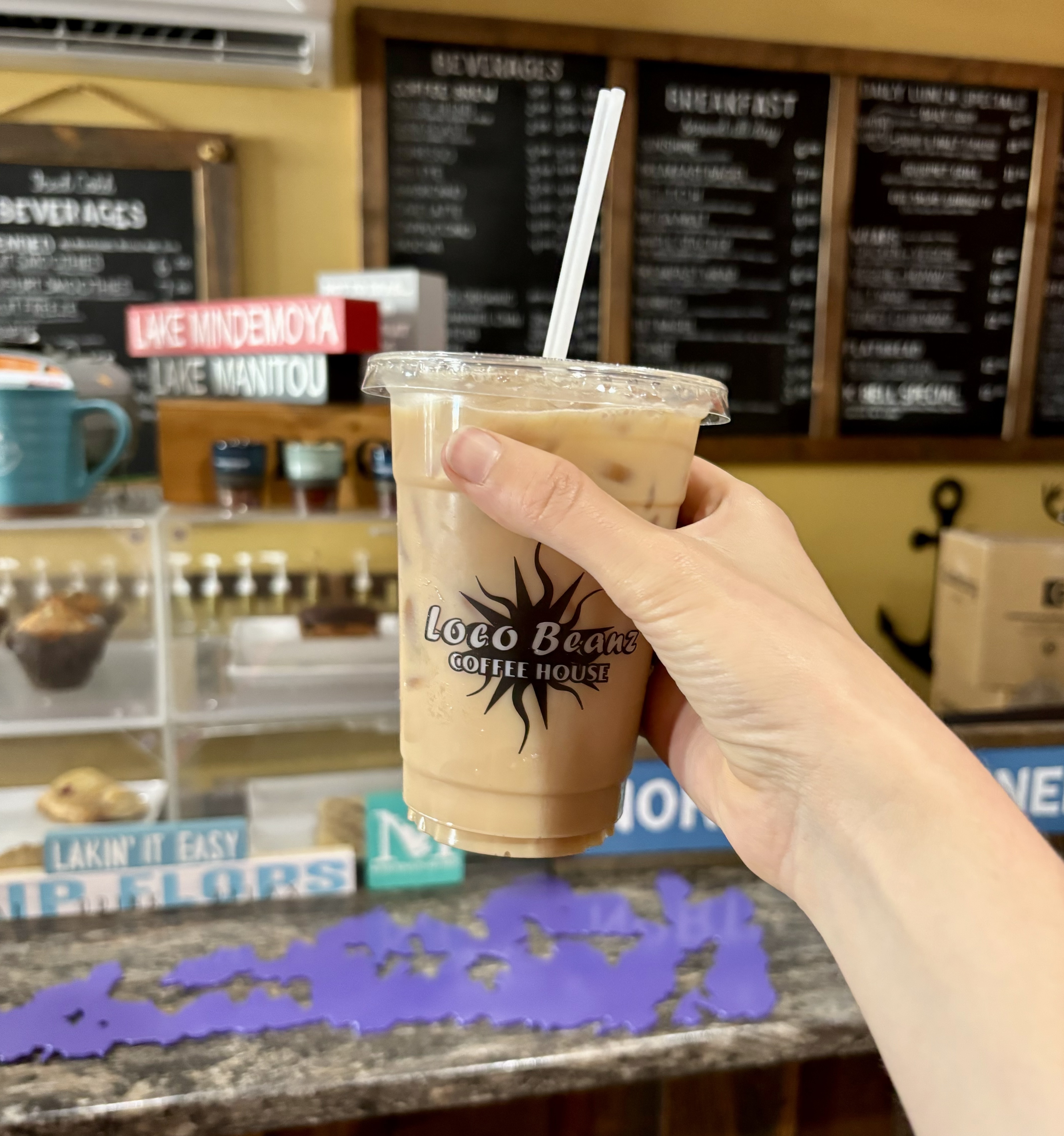 A hand holding up and iced coffee with a Loco Beanz logo on the cup in front of the tidy front counter and menu board.