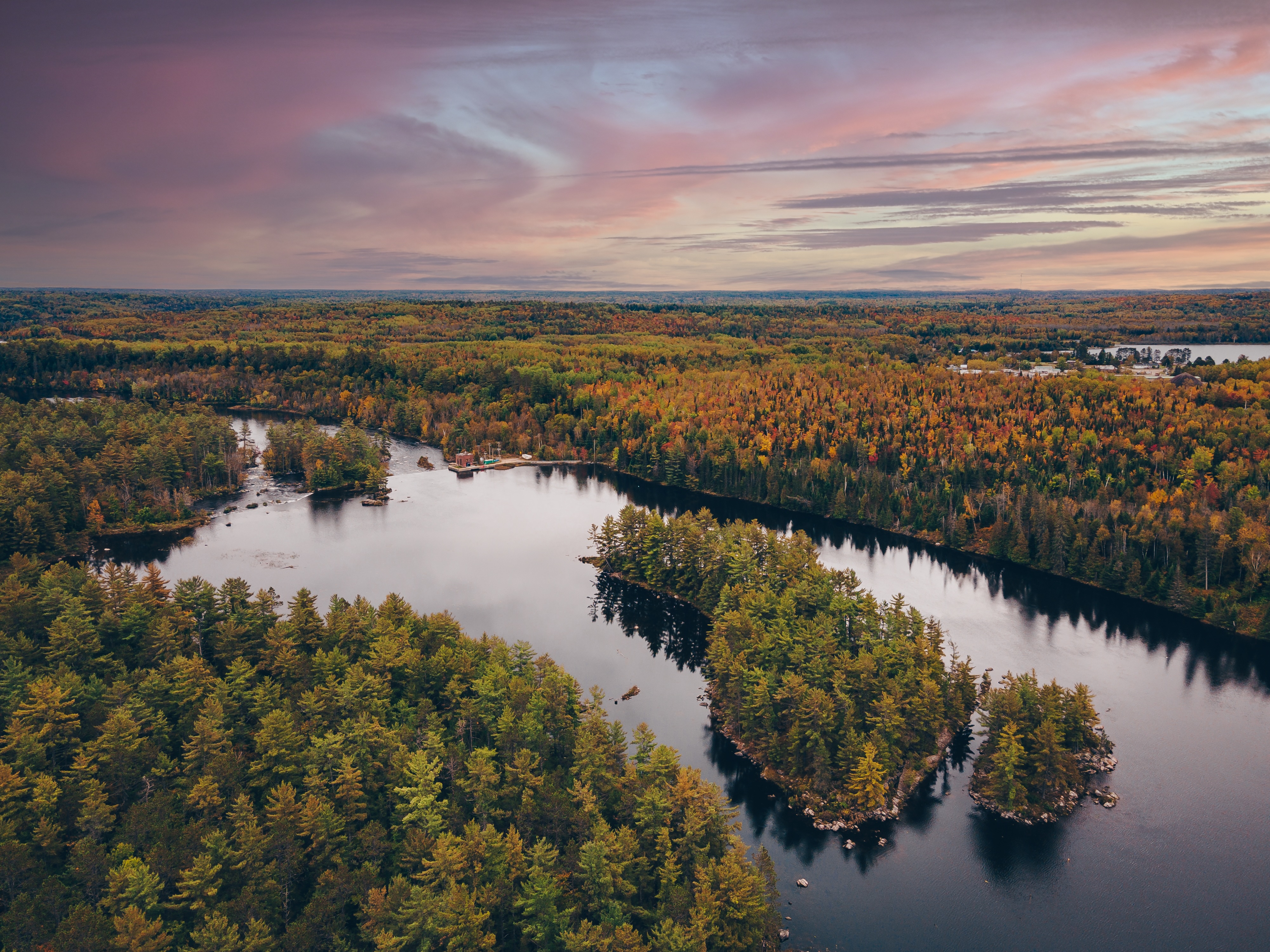 a green forest dotted with changing autumn leaves surrounding a shining lake with a cabin at one end, under swirls of pink clouds at sunset. 