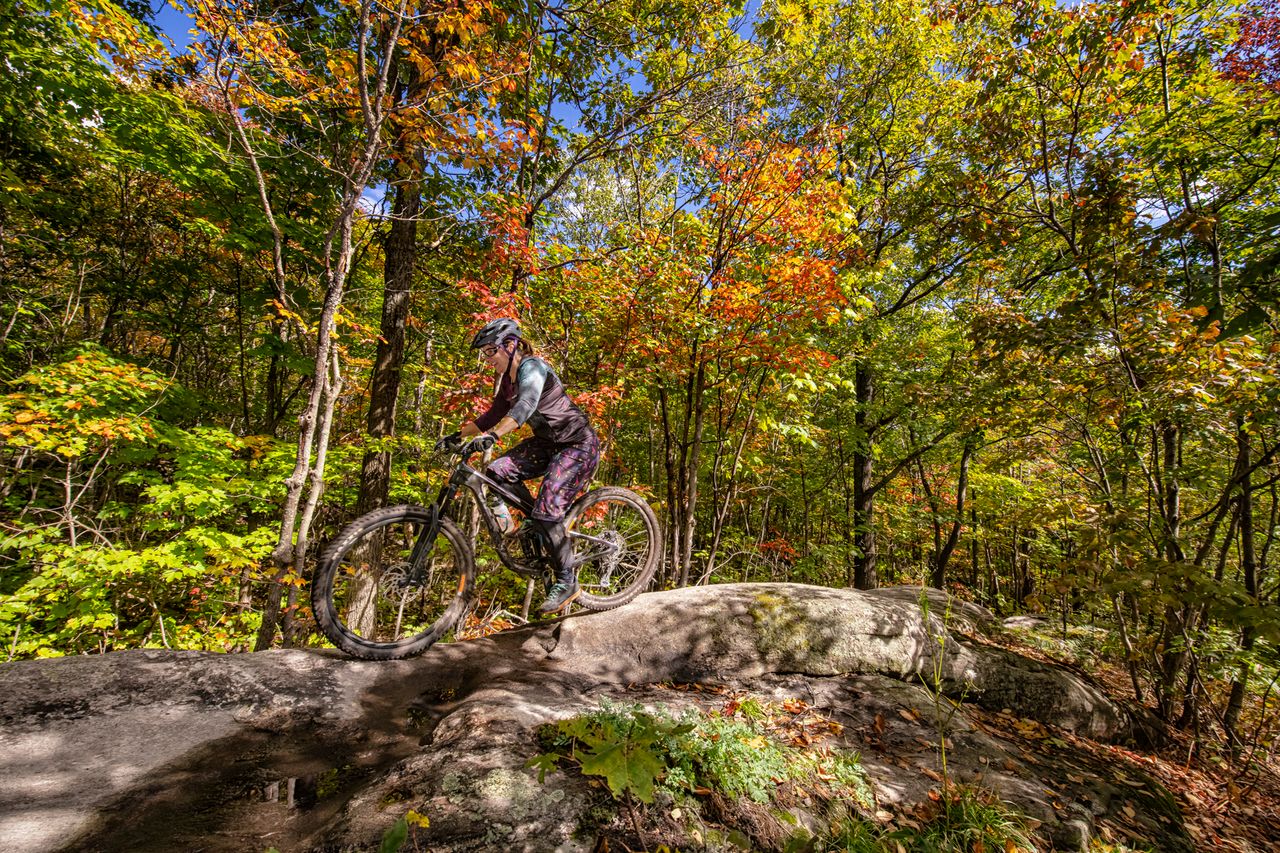 A smiling cyclist rides a mountain bike over a smooth rock section of trail through brightly-coloured autumn forest.