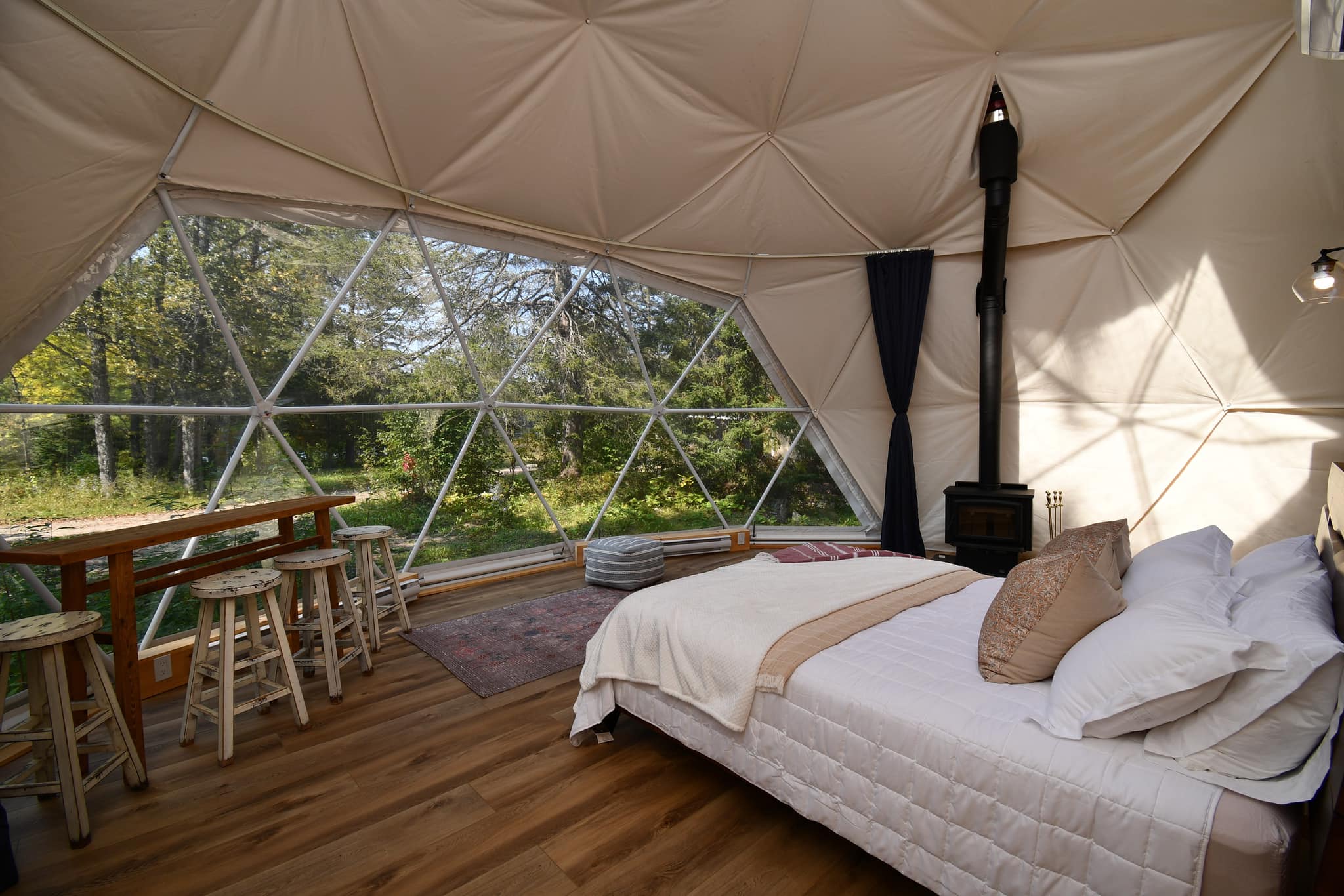 The interior of a glamping dome at Otter Pointe, surrounded by forest outside. It has wooden floors, table and chairs and a white fluffy bed. 