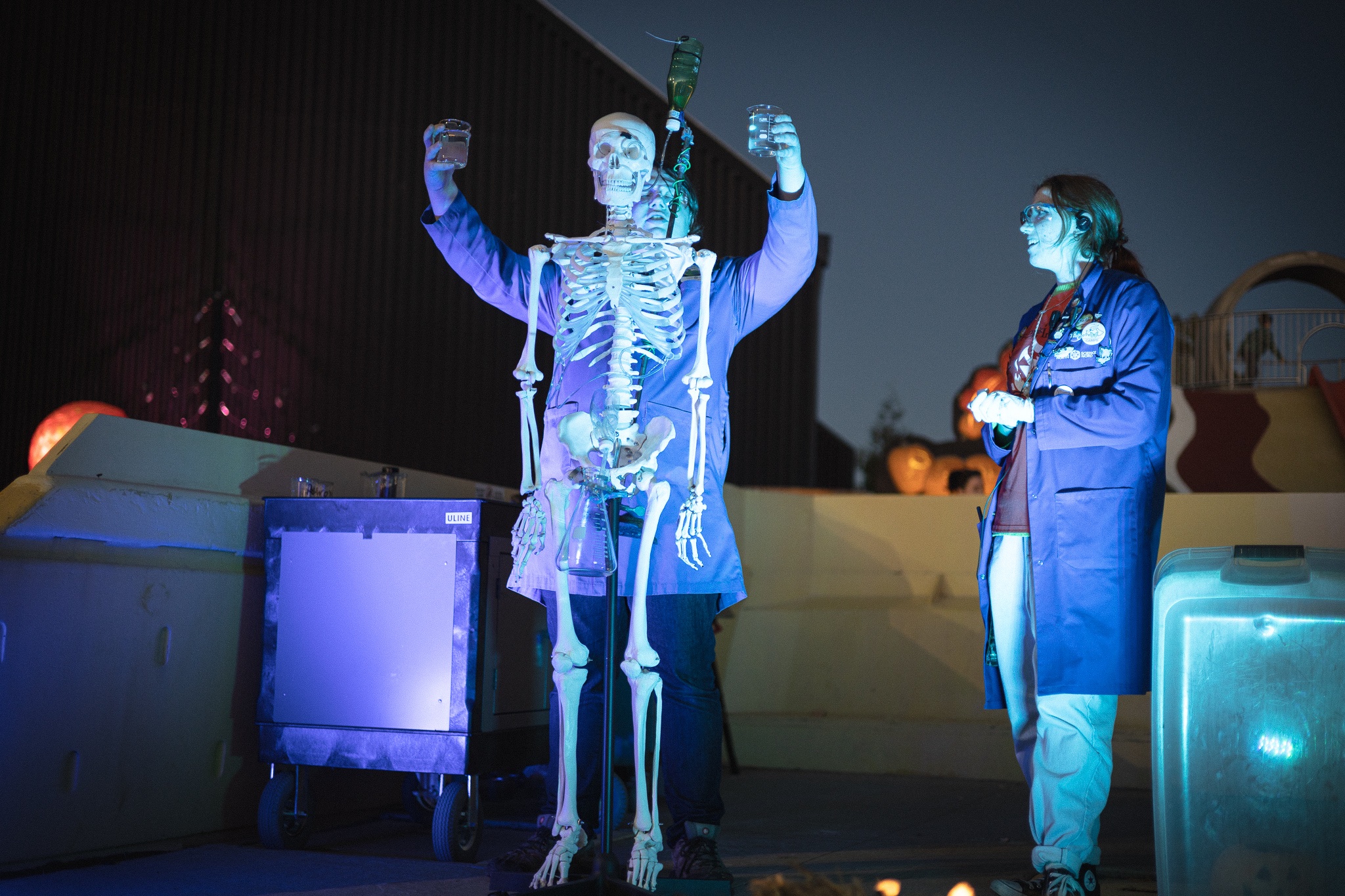 Two scientists from Science North, wearing blue labs coats, stand under spotlights on an outdoor stage at night, performing a science experiment with a skeleton and two beakers of fluid.