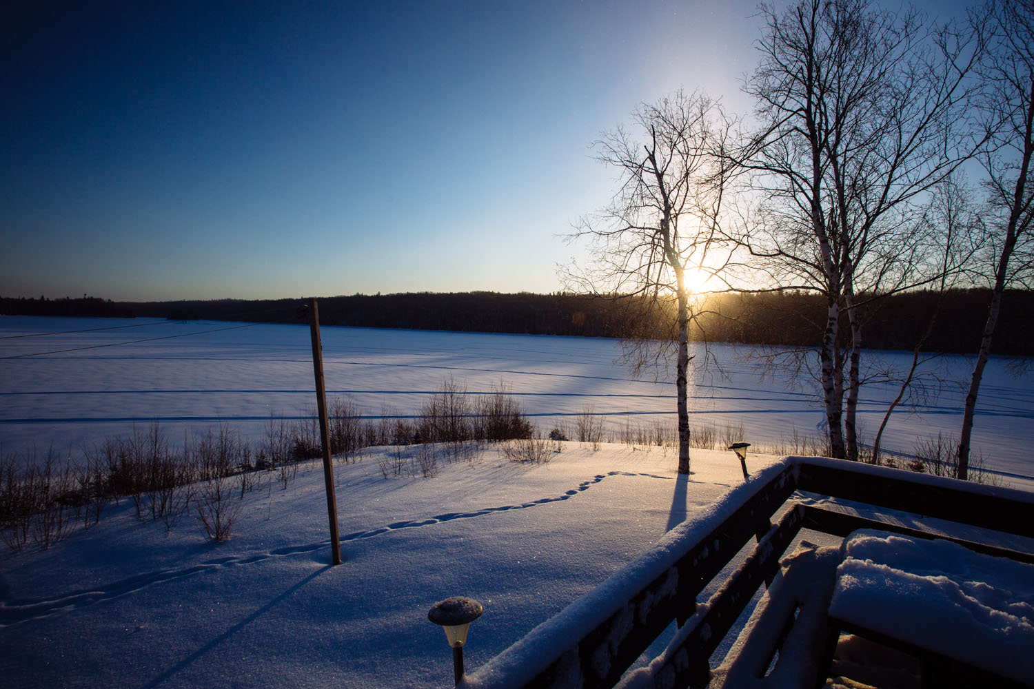 a snowy landscape with a sun setting in the distance behind a tree and the corner of a porch on the bottom right of the image