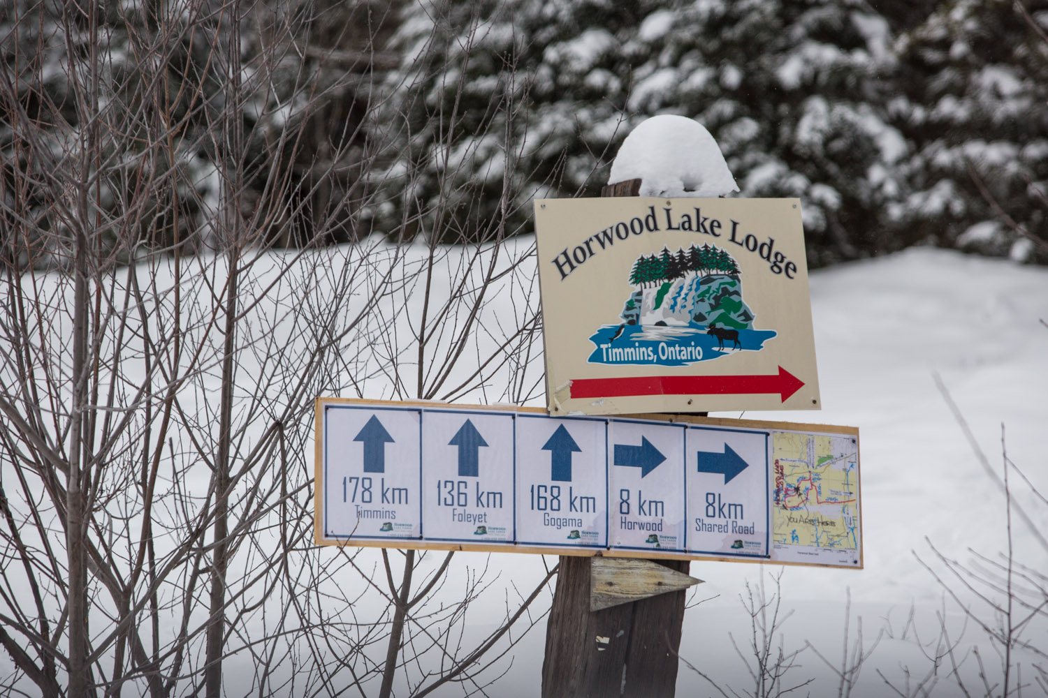 A close-up of a sign that says Horwood Lake Lodge and has an arrow below this and its logo. There are also more arrows stating how far the cities are from it below this