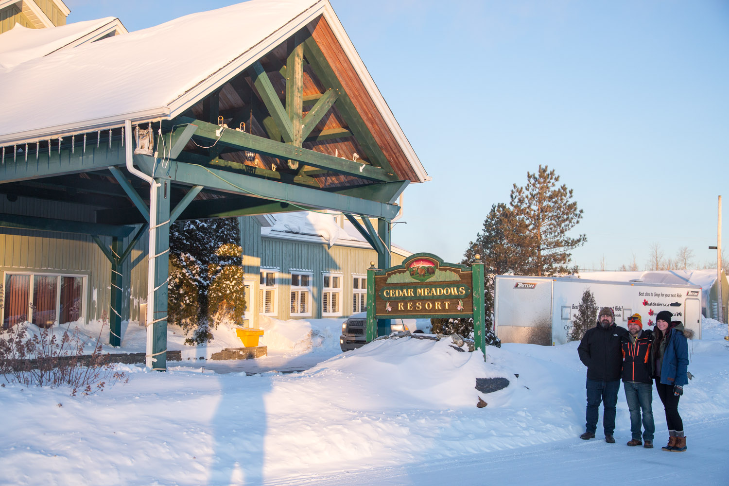 3 people are standing together beside the Cedar Meadows Resort sign which is located right beside the entrance to the building in the wintertime