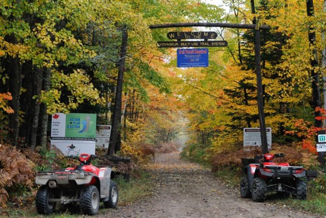 A wooden gateway labelled "Voyageur Multi-Use Trail", opening to a dirt trail surrounded by brightly-coloured autumn forest. ATVs are parked at the gate.