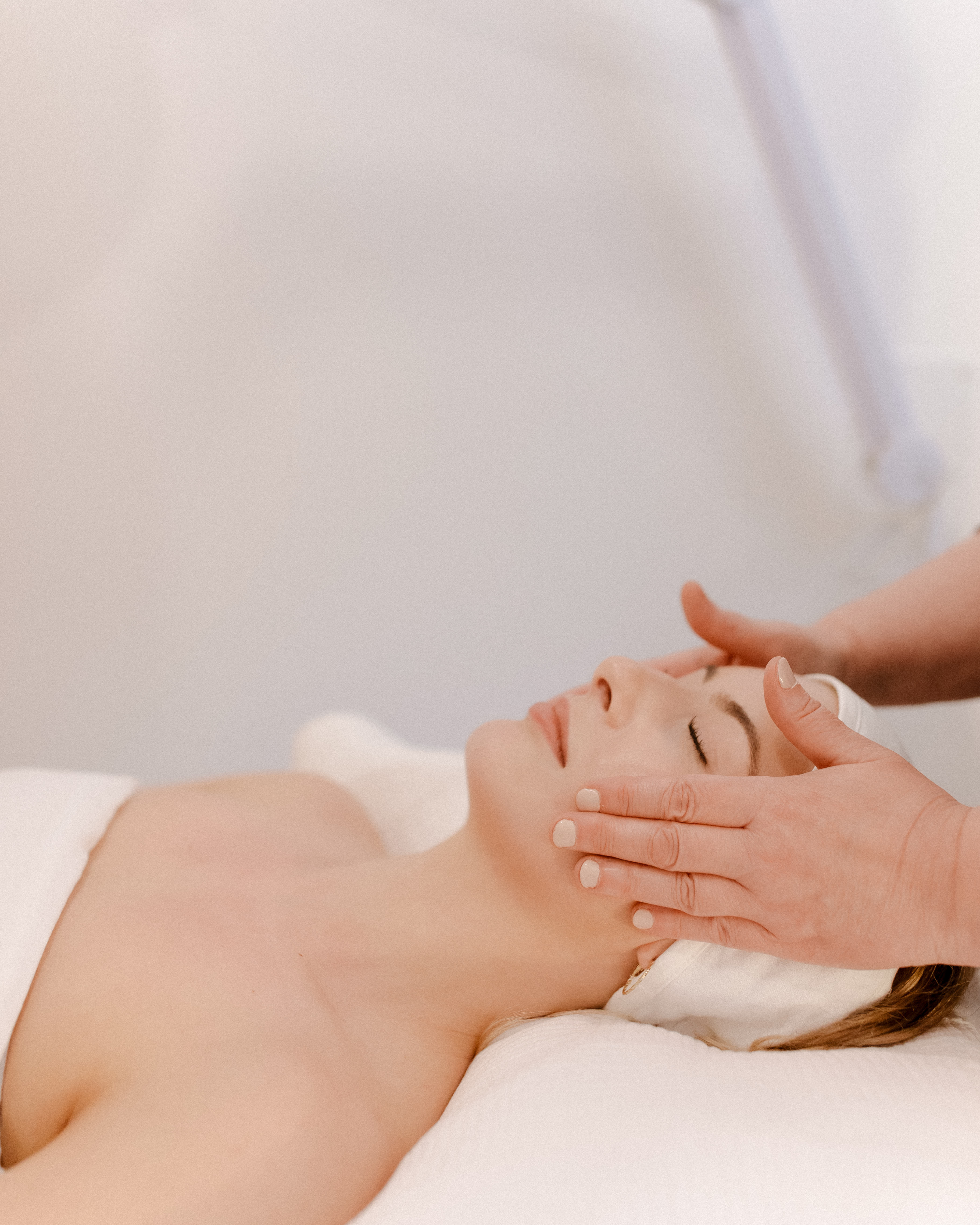 a close-up of the top half of a person who is laying down with their eyes closed and an esthetician's hands are rubbing their face