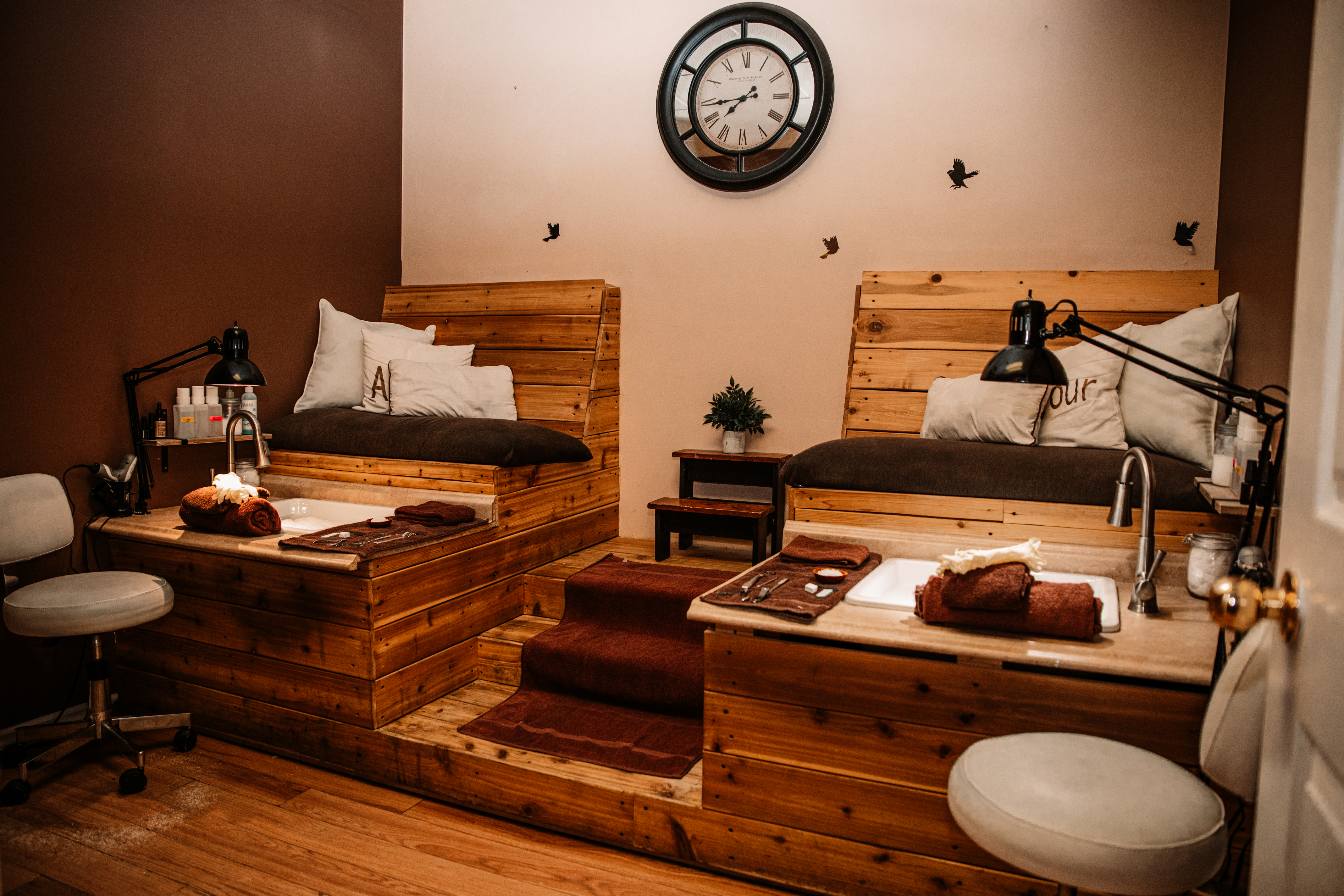 A close-up of the pedicure stations which are made of wood and have lamps on both sides of the image. There is a clock on the white wall in the center of the image.