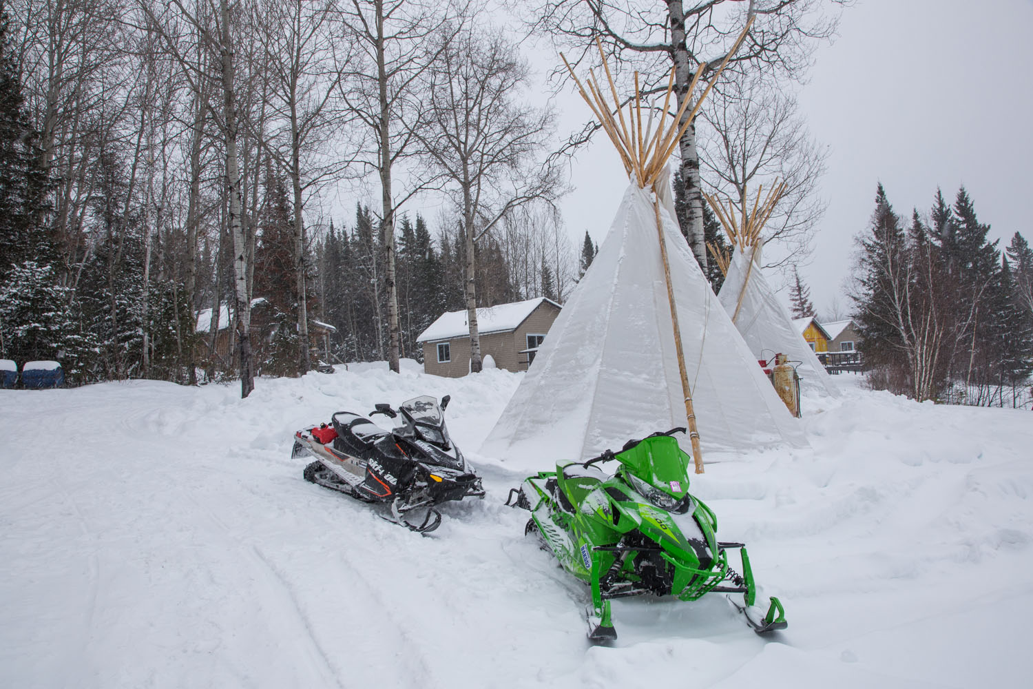 2 snowmobiles 1 black and 1 green are parked beside a tipi. In the distance behind it is another tipi and behind this one you can see the cabins in the distance. There are trees in the background