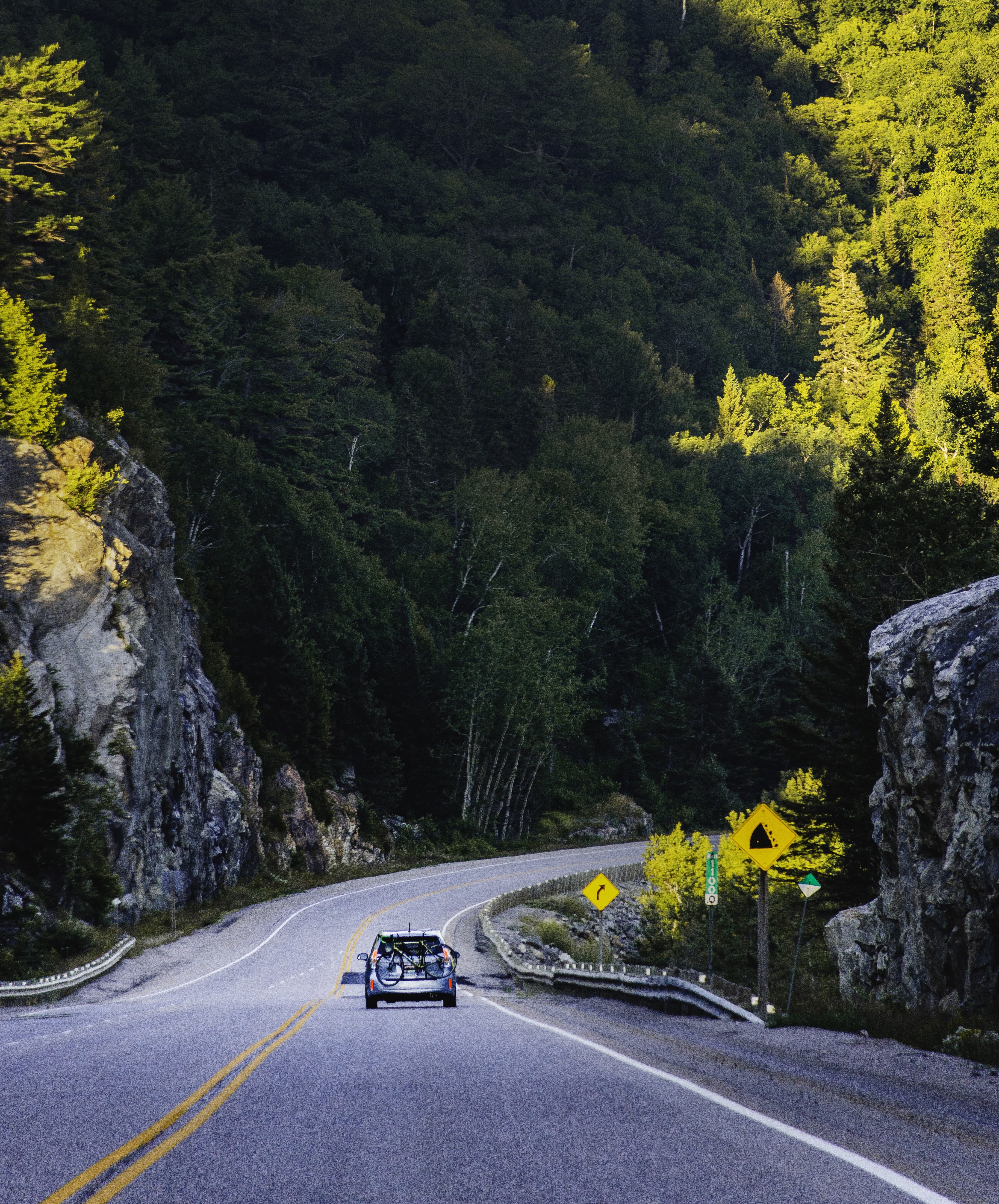 a car drives down a two lane highway with tall rock cliffs covered in forest on either side.