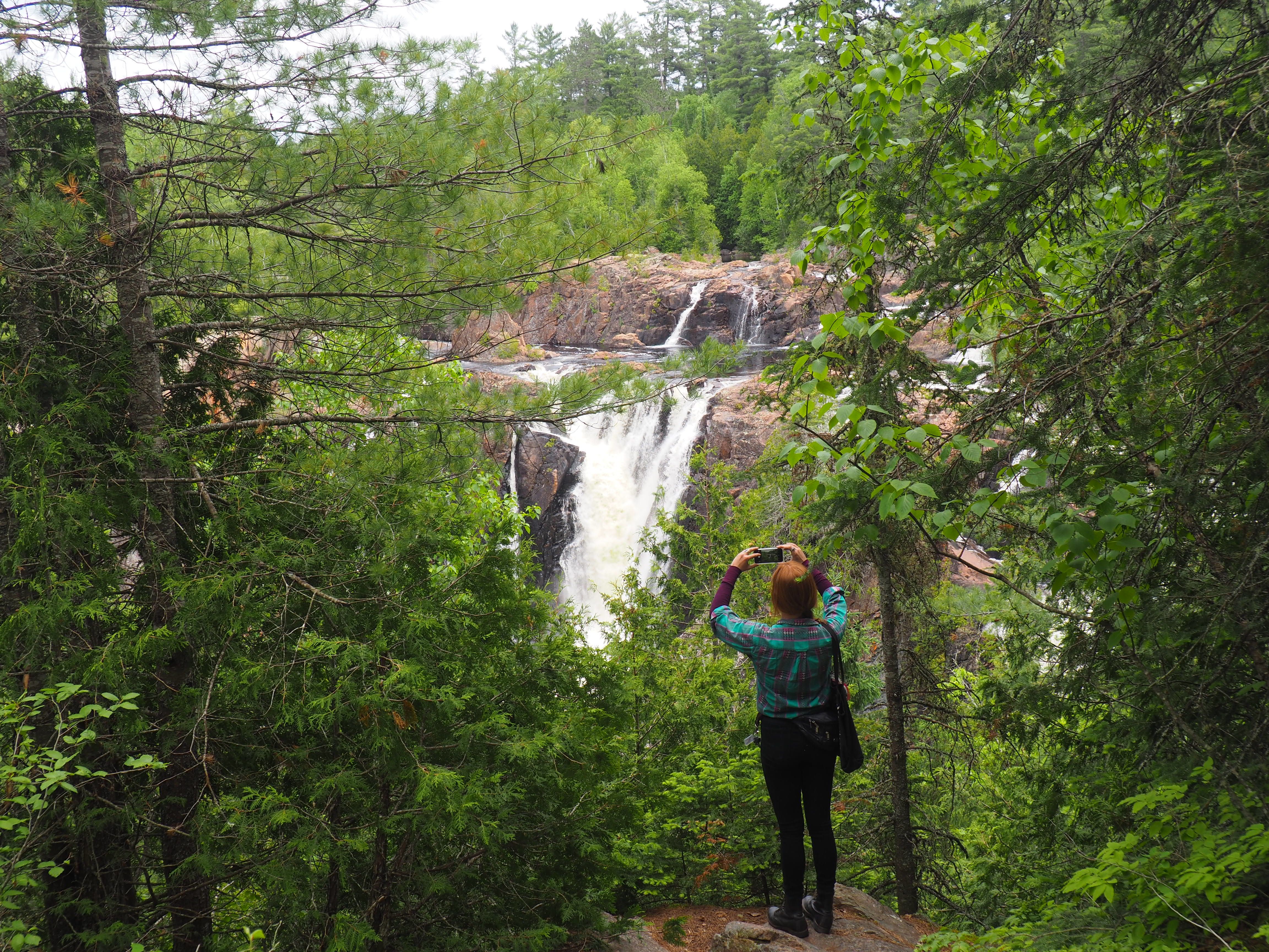 A woman standing in lush green forest snaps a photo of a tall rushing waterfall, Aubrey Falls.