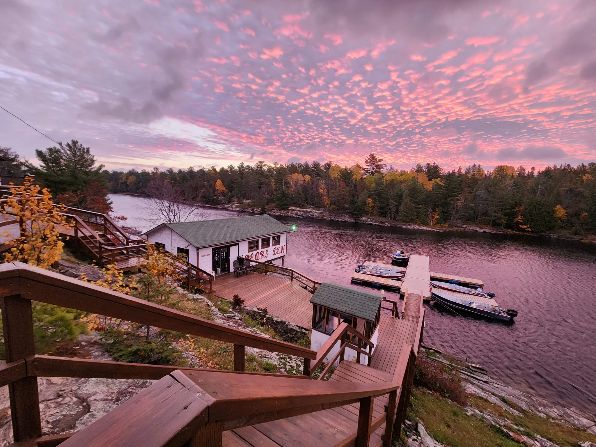 Bear's Den Lodge; a wooden staircase leading down a riverbank to a dock, next to a white and green lodge on the shore. It is surrounded by red, green and yellow autumn forest under a pink sunrise.