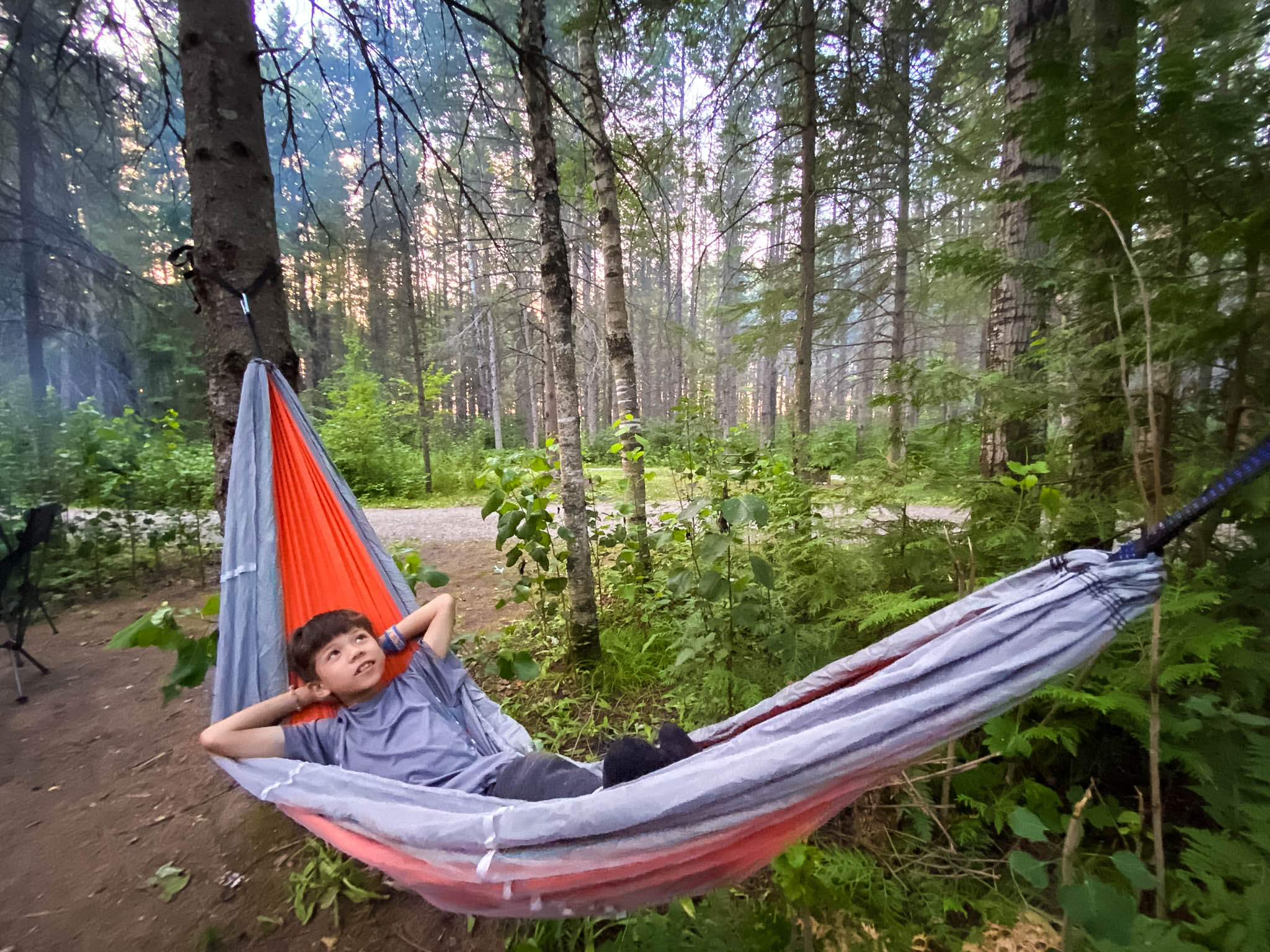 a boy lays in a hammock strung between two trees in a forested campsite. He has his hands clasped behind his head as he looks contentedly up at the trees. 