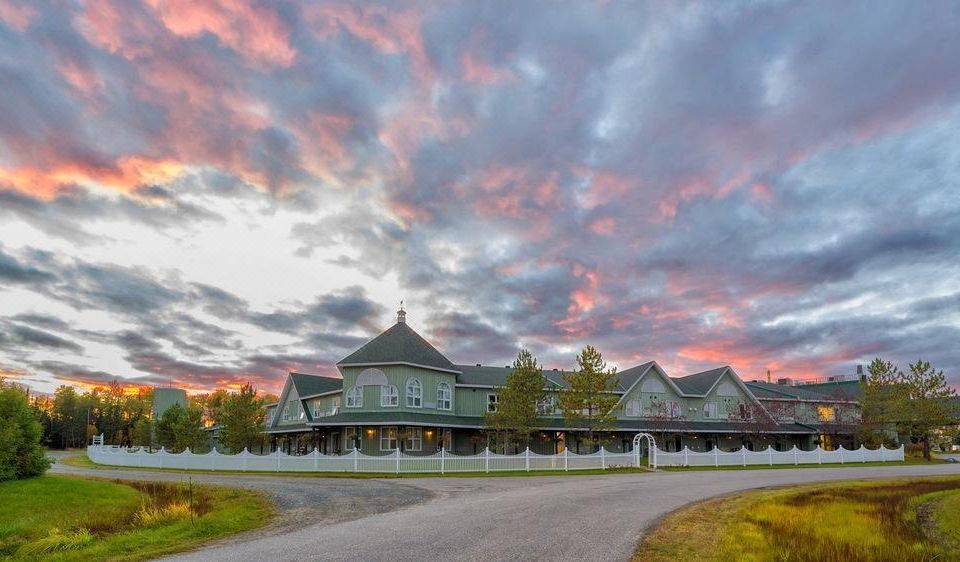 Cedar Meadows Spa; a sprawling mansion resort with many peaked roofs and a white picket fence, surrounded in autumn trees under a pink and purple sunset. 