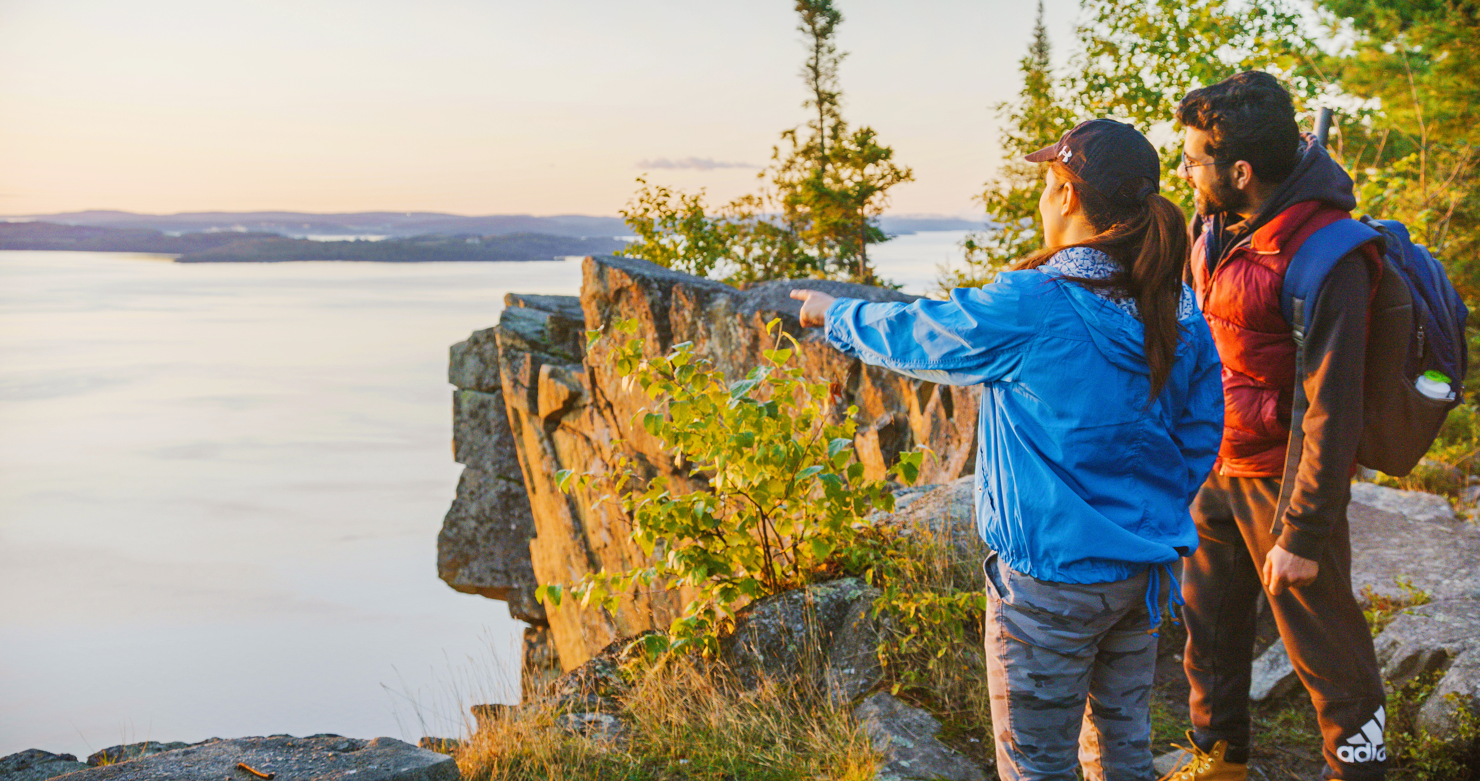 two hikers stand at the top of Devil's Rock, a tall cliff overlooking Lake Temiskaming, surrounded in green forest.