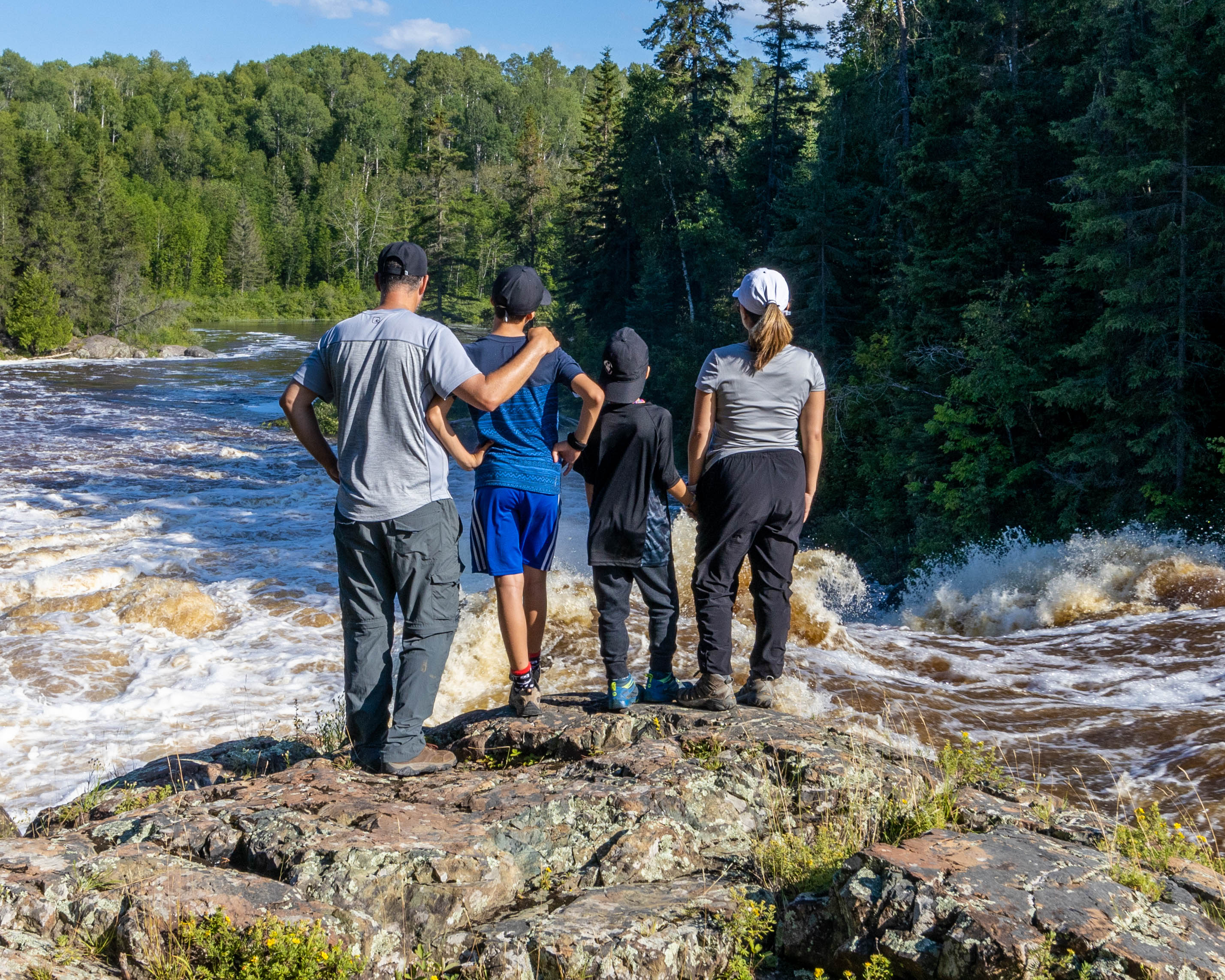 A family of four stands side by side on a rock bank looking at the whitewater river surrounded by green forest on the Hell's Gate Trail.